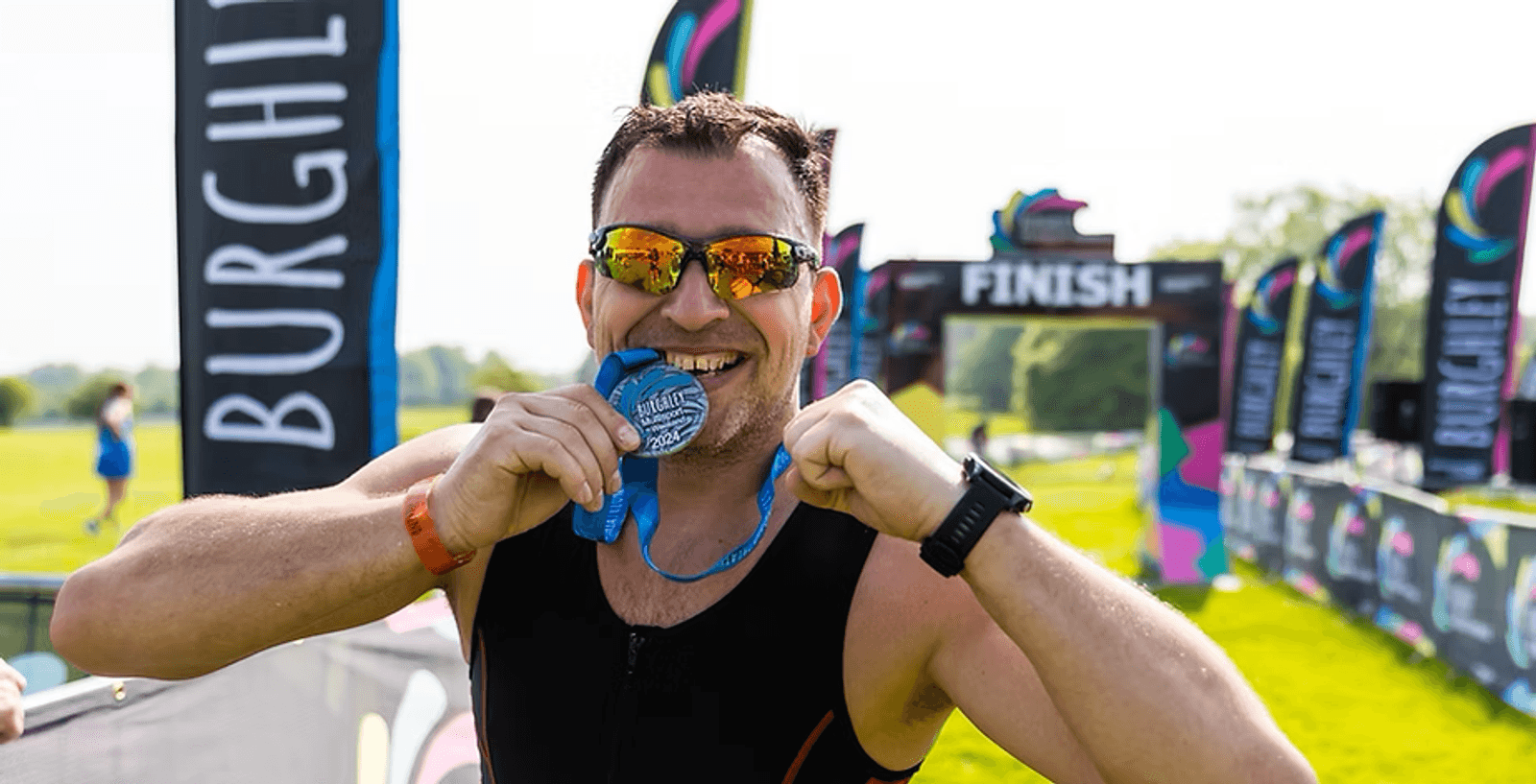 A man wearing sunglasses and a sports outfit bites a medal and smiles at a finish line, surrounded by banners. It's a sunny day and the atmosphere appears festive and celebratory.