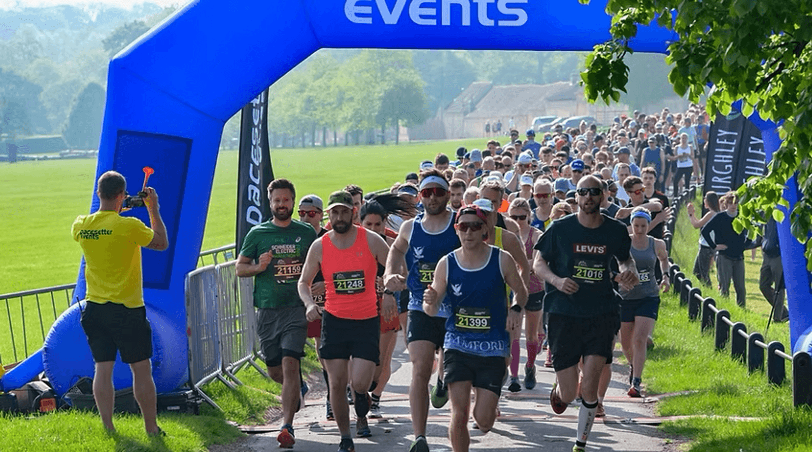 A group of runners in athletic gear starting a race under a blue inflatable arch in a park. The path is lined with spectators and green trees, and a sunny sky is overhead. A volunteer in a yellow shirt is standing nearby.