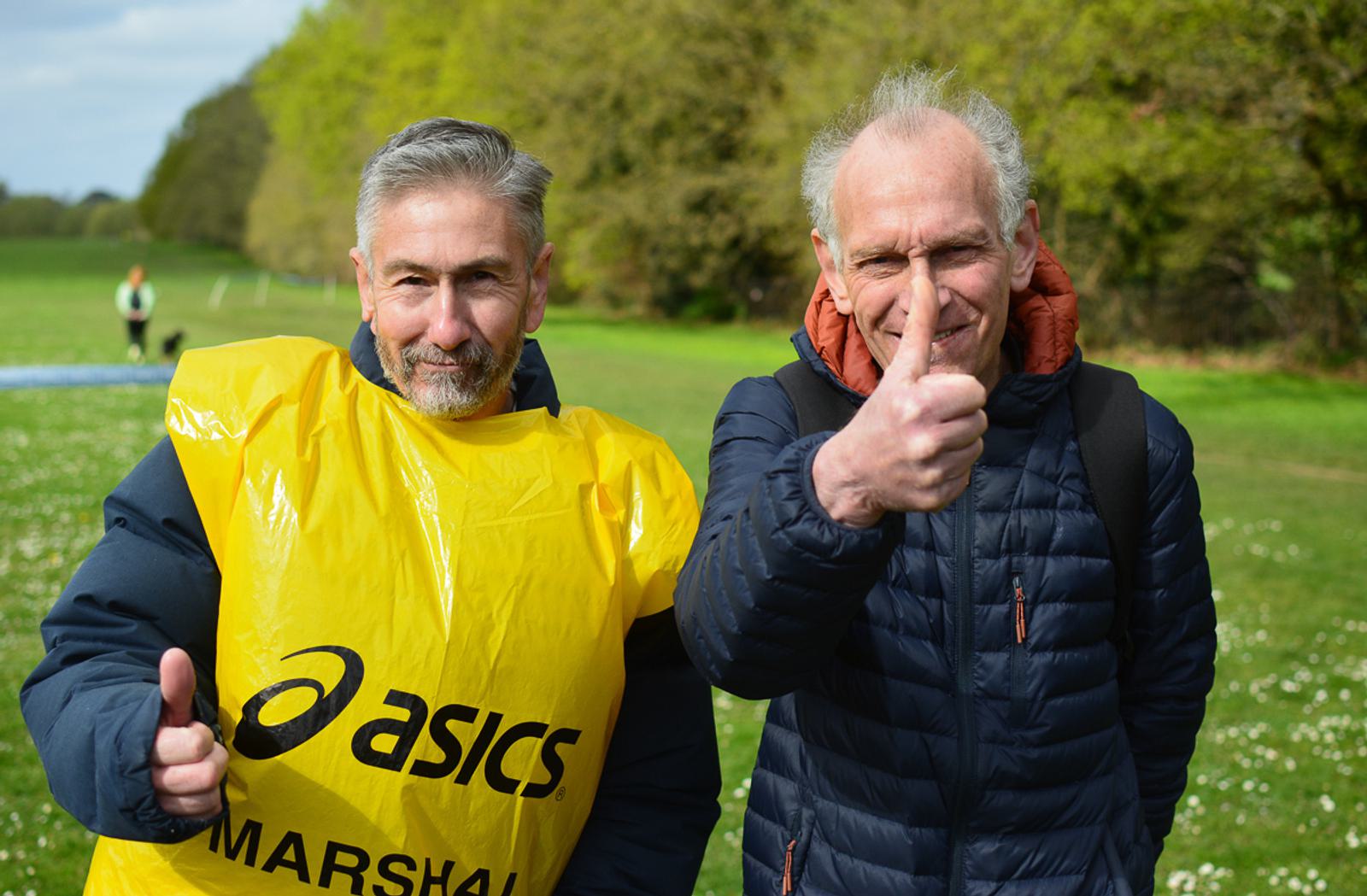 Two men standing outside on a grassy field, both giving thumbs-up. One wears a yellow Asics marshal vest, and the other is in a blue quilted jacket. Trees line the background under a partly cloudy sky.