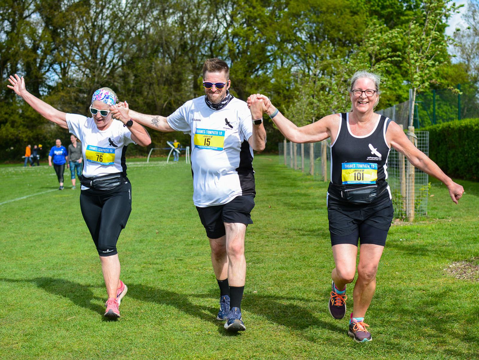 Three people in sports attire hold hands and raise their arms triumphantly as they run on a grassy field. They are smiling, wearing numbered bibs, and surrounded by trees and additional runners in the background.