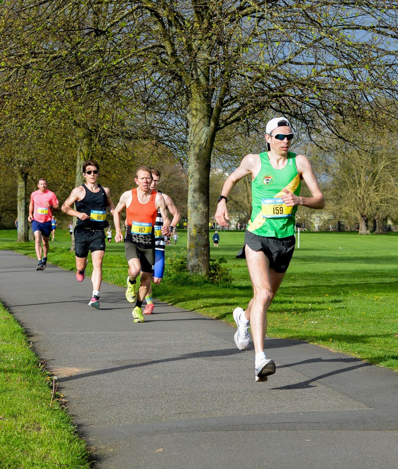 Four runners are participating in an outdoor race on a paved path through a green park. The front runner wears a green top and sunglasses, while the others follow closely. Trees line the path, and the sky is clear.