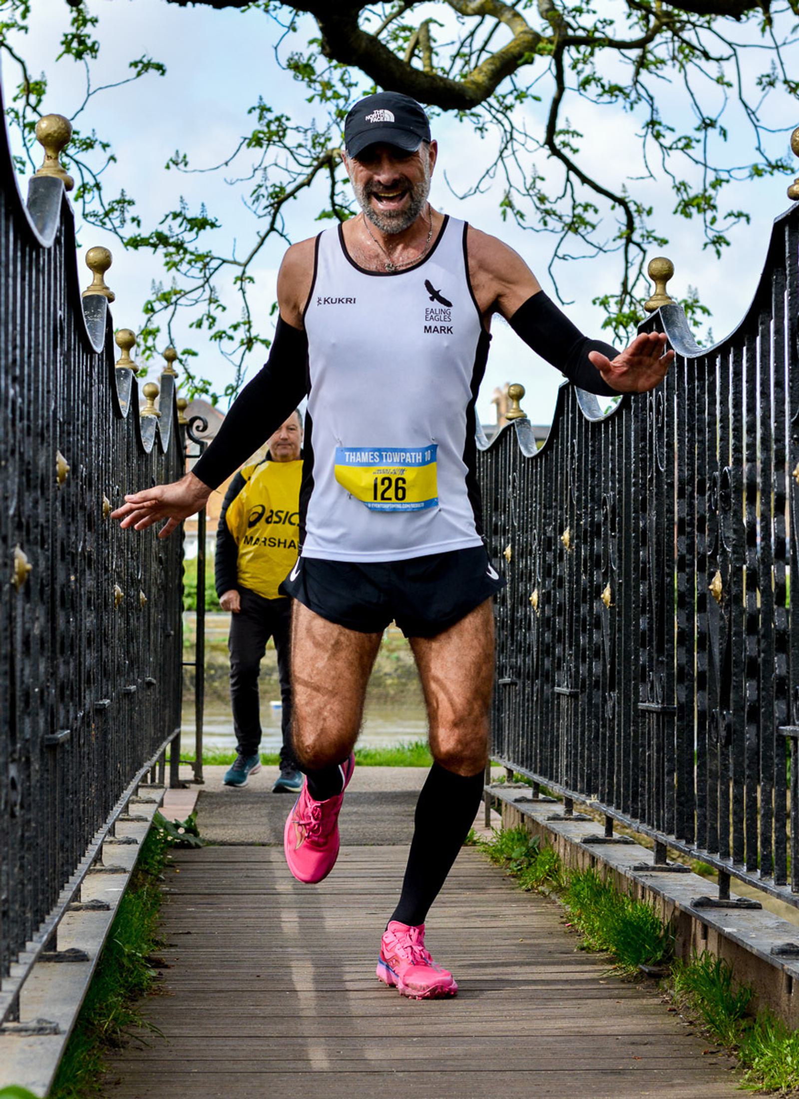 A male runner in pink shoes and a white tank top with "#126" approaches the finish line on a bridge, smiling and spreading his arms. Another person follows behind in a yellow shirt. The bridge has ornate metal railings and a tree in the background.