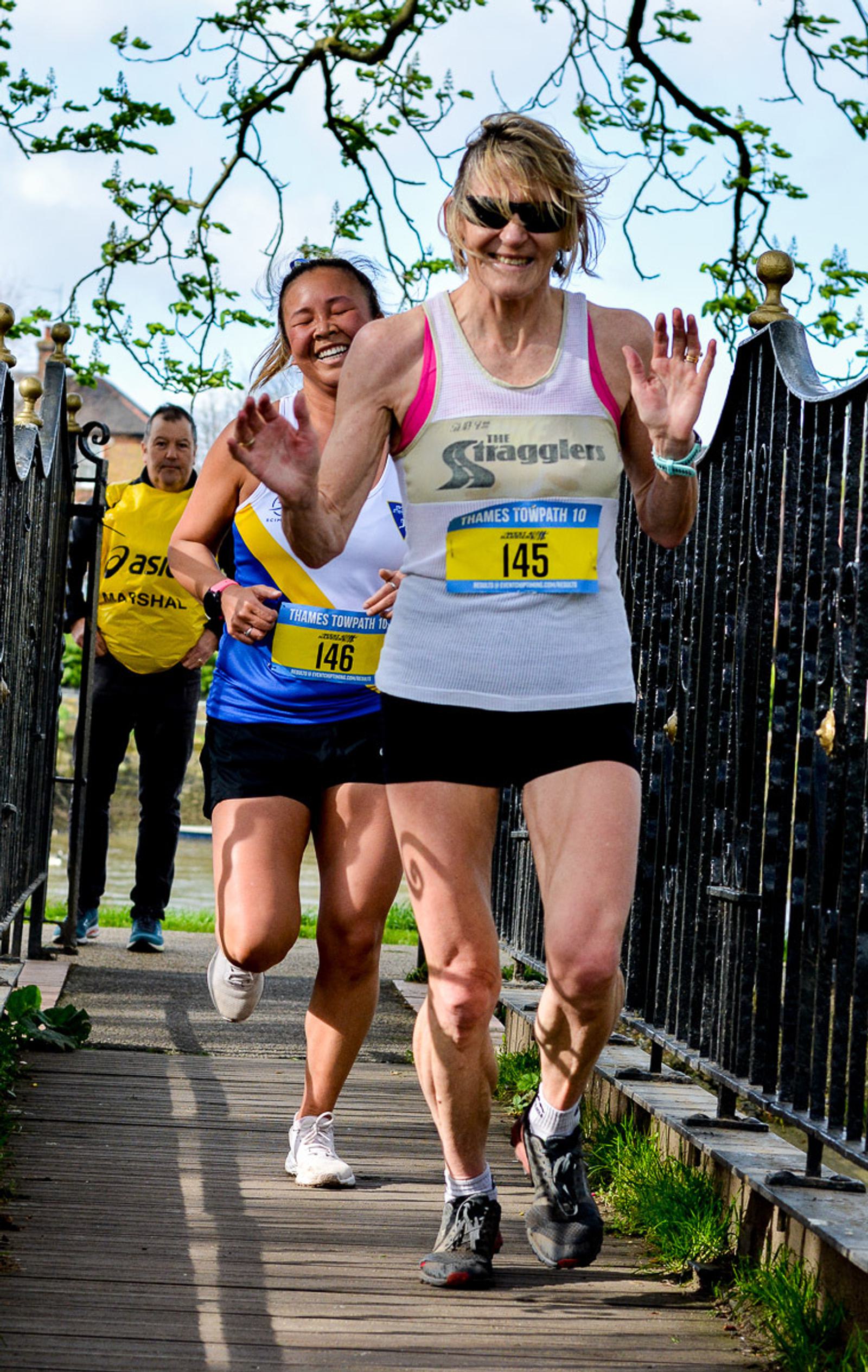 Two women smiling and running in a race, wearing bib numbers 145 and 146. They are on a pathway with metal railings and a third person is running behind them. The background has trees and a blue sky.