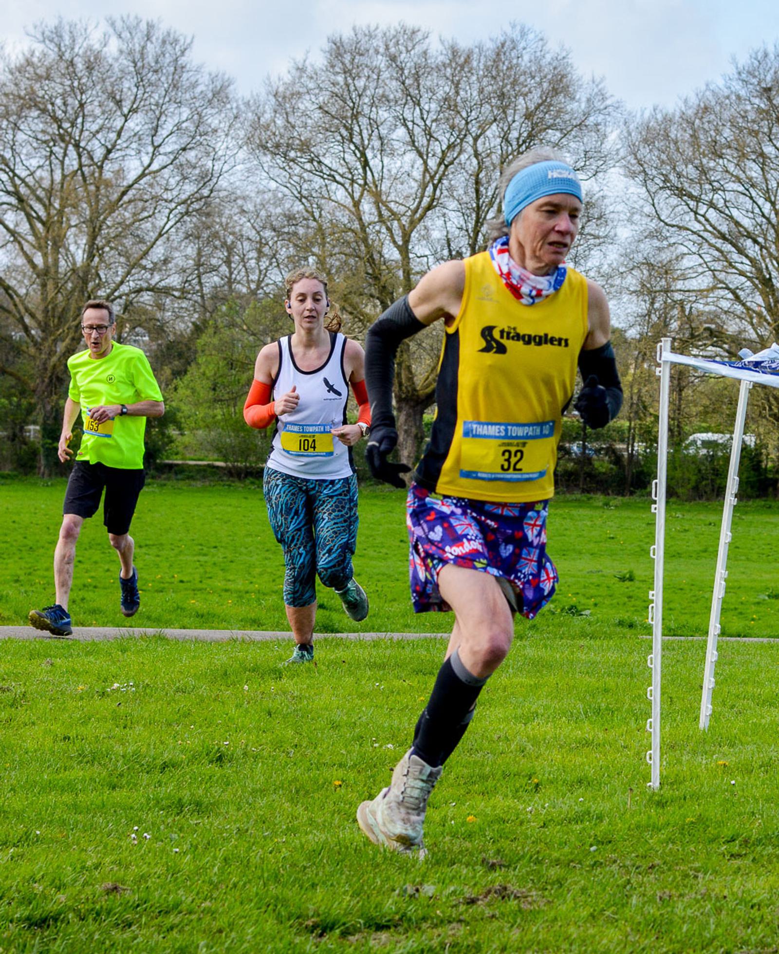 Three runners in a cross-country race on a grassy field. Foreground runner wears a yellow tank and colorful shorts. Behind, a woman in a white tank and a man in a bright green shirt follow. Leafless trees and a cloudy sky in the background.