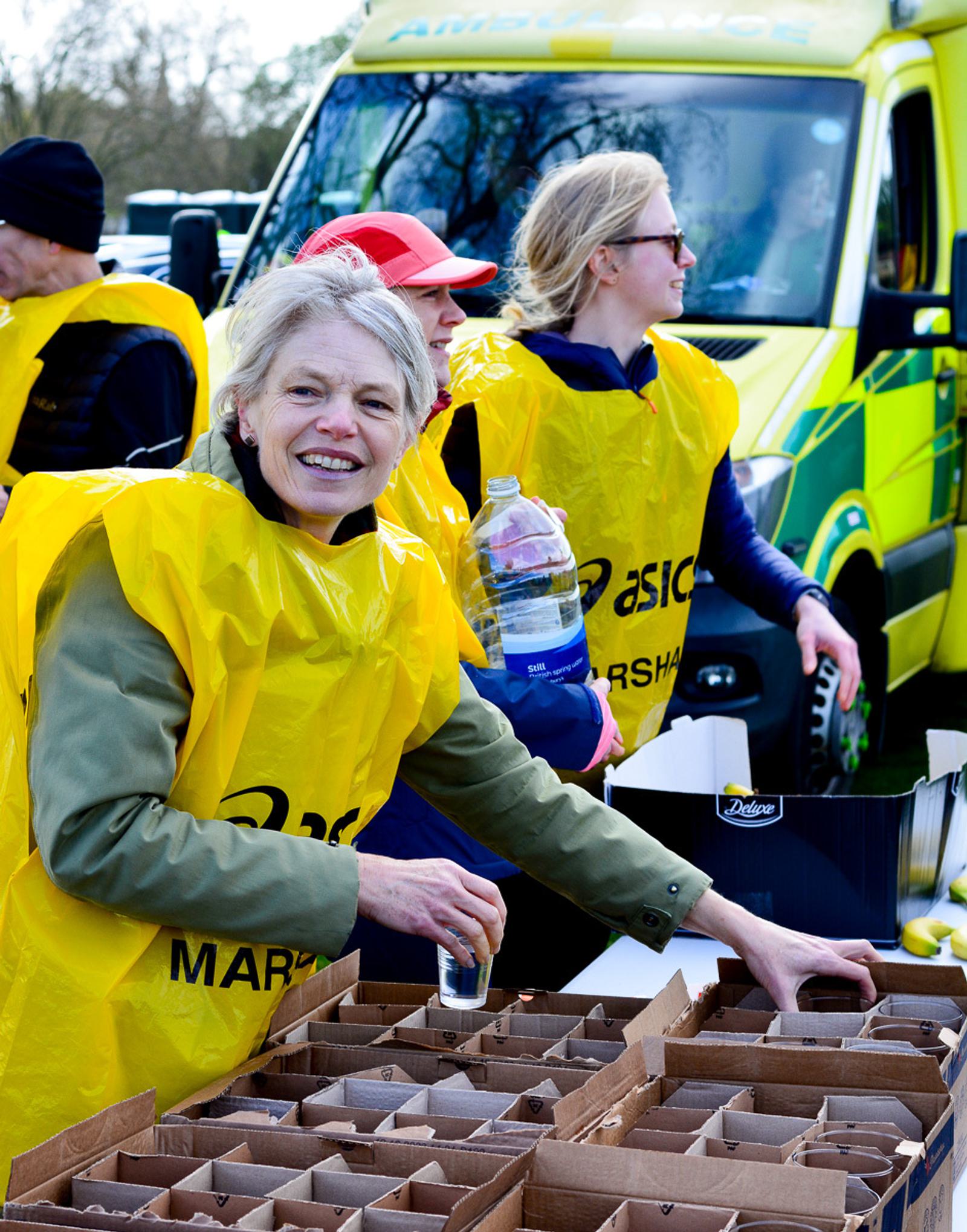 Volunteers wearing yellow vests distribute water bottles at an outdoor event. They stand by a table with cardboard boxes in front of a green and yellow vehicle. One volunteer smiles at the camera while organizing the bottles.