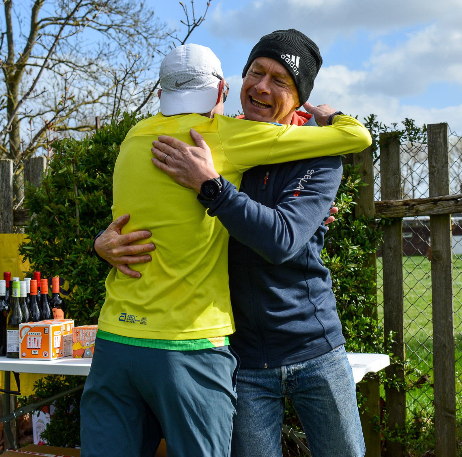 Two people embrace outdoors beside a table with drinks. One wears a yellow jacket and white cap, the other wears a blue jacket and black beanie. A wooden fence and trees are in the background.