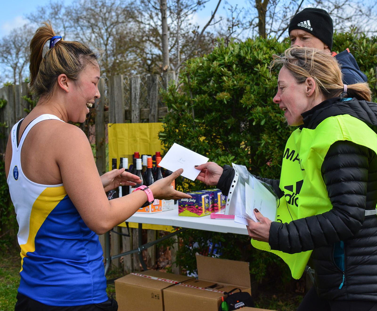 Two women exchange papers at an outdoor event. One wears a running outfit, smiling, while the other, in a safety vest, holds envelopes. In the background, there are drinks, snacks, and trees, with a couple of spectators nearby.