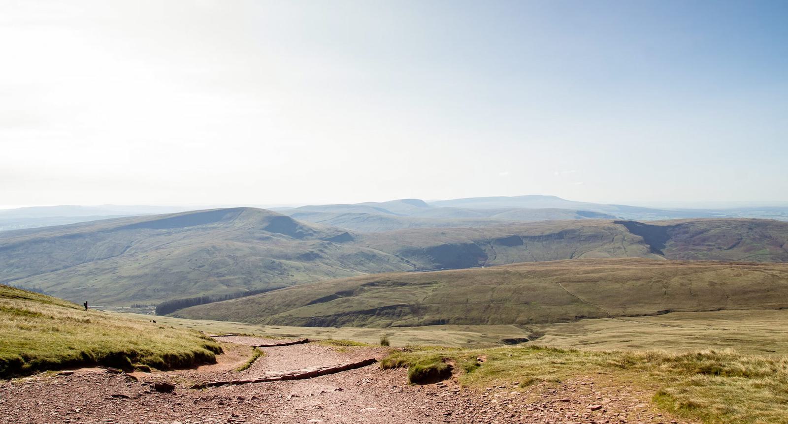 A scenic view of a mountain landscape with rolling hills under a clear blue sky. A dirt path winds through grassy terrain, leading towards distant peaks.