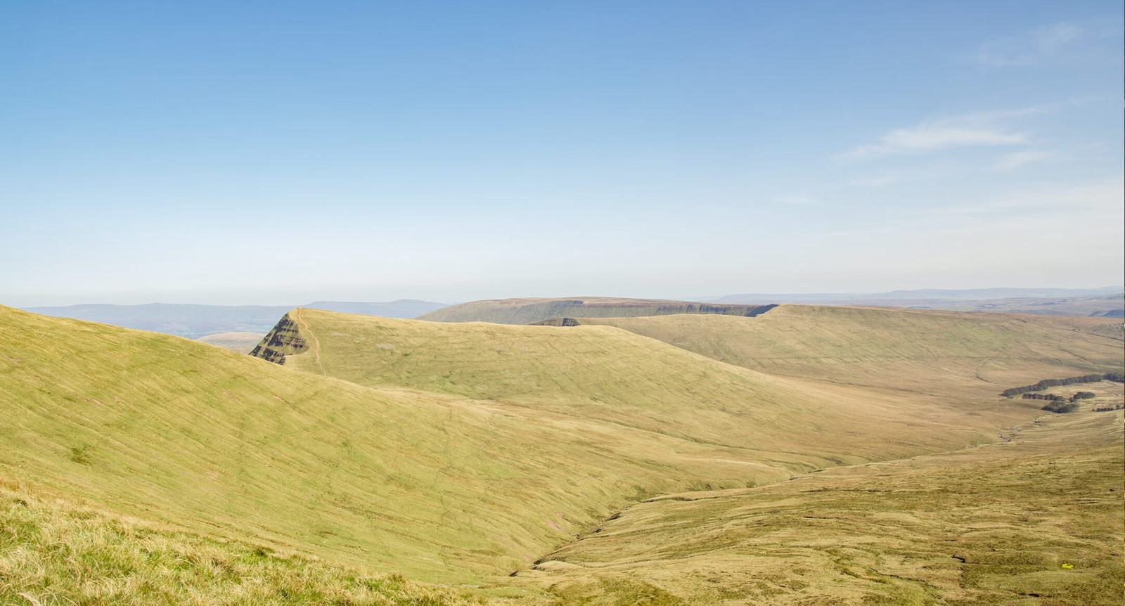 A wide landscape view of rolling green hills under a clear blue sky, with a single prominent hill in the distance. The terrain is grassy with occasional patches of brown and yellow, suggesting a transitional season.