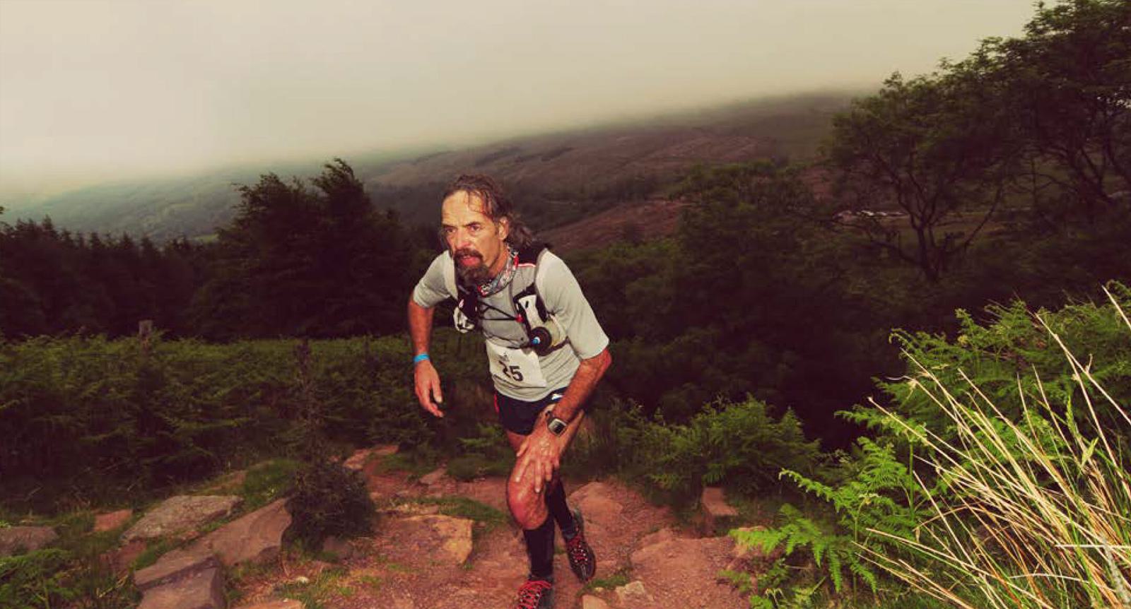 A man in athletic gear and a race number runs uphill on a rocky trail surrounded by lush greenery. The sky is overcast, and the landscape in the background is blurred by mist and fog.