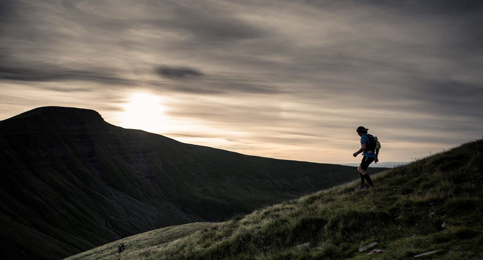 A person wearing a backpack hikes along a grassy hillside at dusk. The sun sets behind distant mountains, casting dramatic shadows across the landscape. The sky is filled with wispy clouds, creating a serene and adventurous atmosphere.