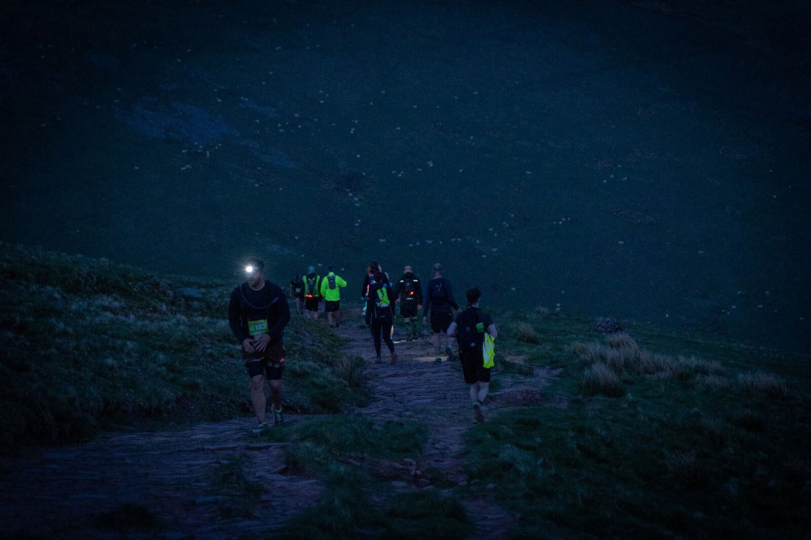 A group of people hiking on a dimly lit trail at dawn or dusk. One hiker has a headlamp, illuminating part of the pathway. The landscape is grassy and hilly, creating a serene, early morning or evening atmosphere.