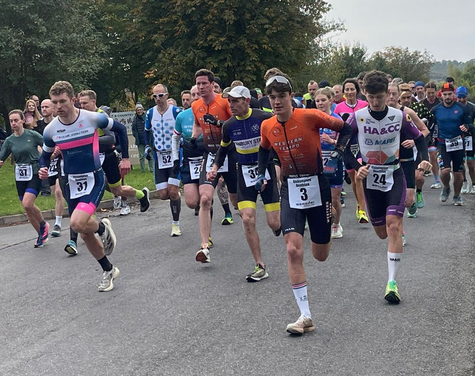 A group of runners at the start of a race, wearing athletic gear and numbered bibs. They are running on a paved road with trees and spectators in the background.