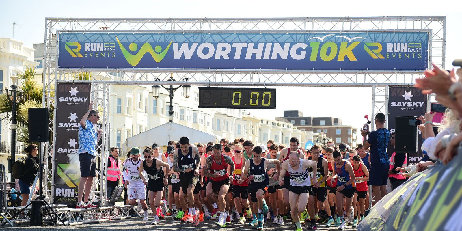 Runners at the starting line of the Worthing 10K race. A large crowd of participants, wearing athletic gear, is poised to begin the race beneath a banner displaying "WORTHING 10K." Spectators stand on the sidelines. The timer above shows 0:00.