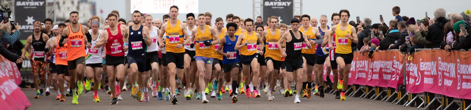 A large group of runners in colorful athletic gear start a race on a road lined with cheering spectators behind pink barricades on a cloudy day.