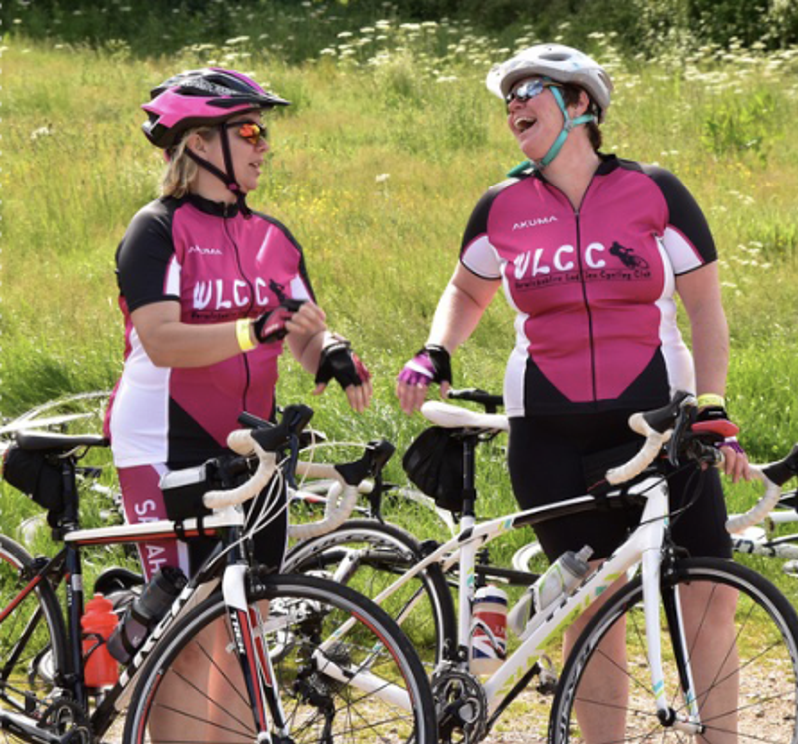 Two women wearing matching pink cycling jerseys stand beside their bikes, smiling and talking. They are both wearing helmets and sunglasses. The background is a grassy area with wildflowers.