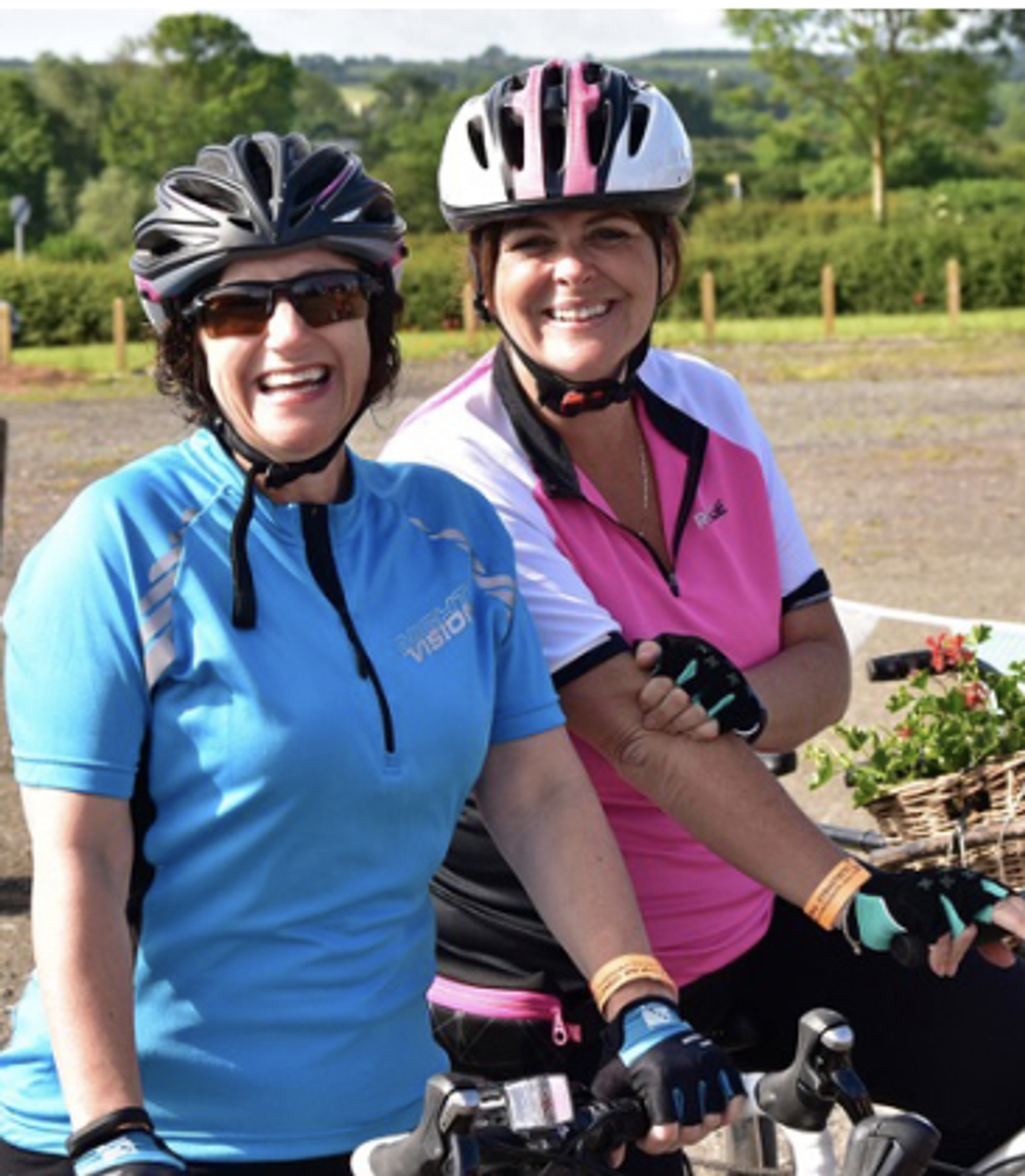 Two women in cycling gear, one in a blue jersey and the other in a pink one, smile while leaning on their bicycles. Both wear helmets and gloves. A basket of flowers is visible on one bike. A scenic, green background enhances the outdoor setting.