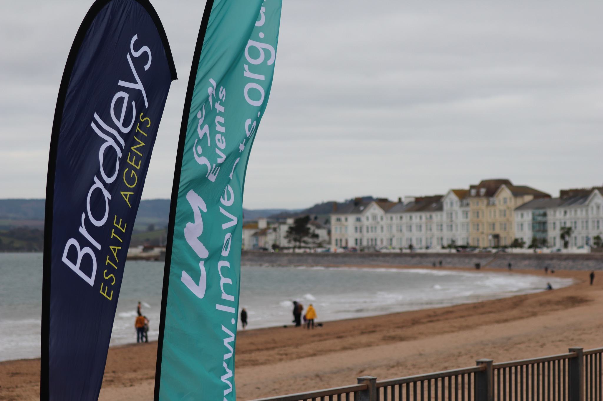 Two promotional flags on a beach; one reads "Bradleys Estate Agents" and the other displays a website URL. People walk along the shore, with seaside buildings and a hilly landscape in the background under a cloudy sky.