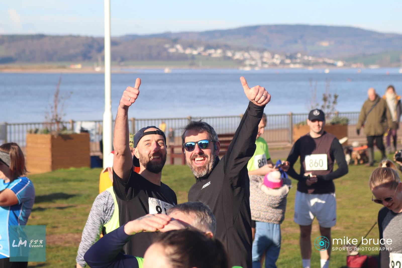 Two men in athletic gear smile and give thumbs up to the camera. They stand outdoors near a body of water, with other runners and participants in the background. One man wears sunglasses, and both display event numbers on their shirts.