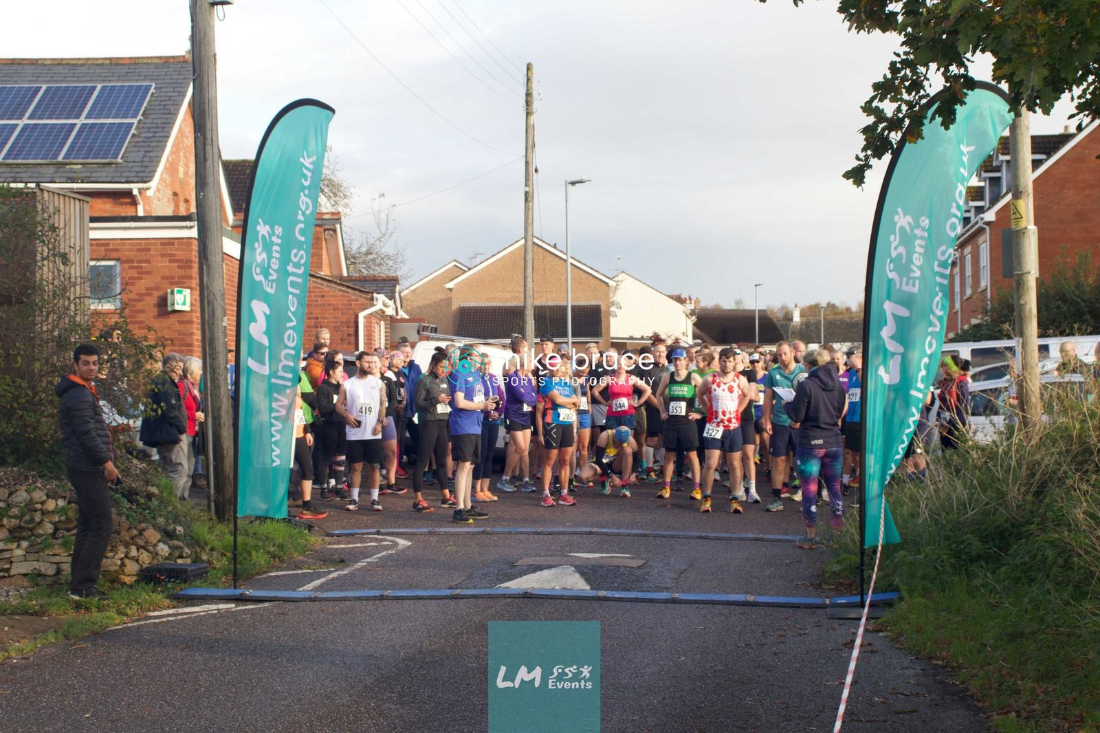 A group of runners gather at the starting line for a race, with LM Events banners on either side. Residential houses and trees are in the background, and a few people are standing nearby, including a person in an orange jacket.