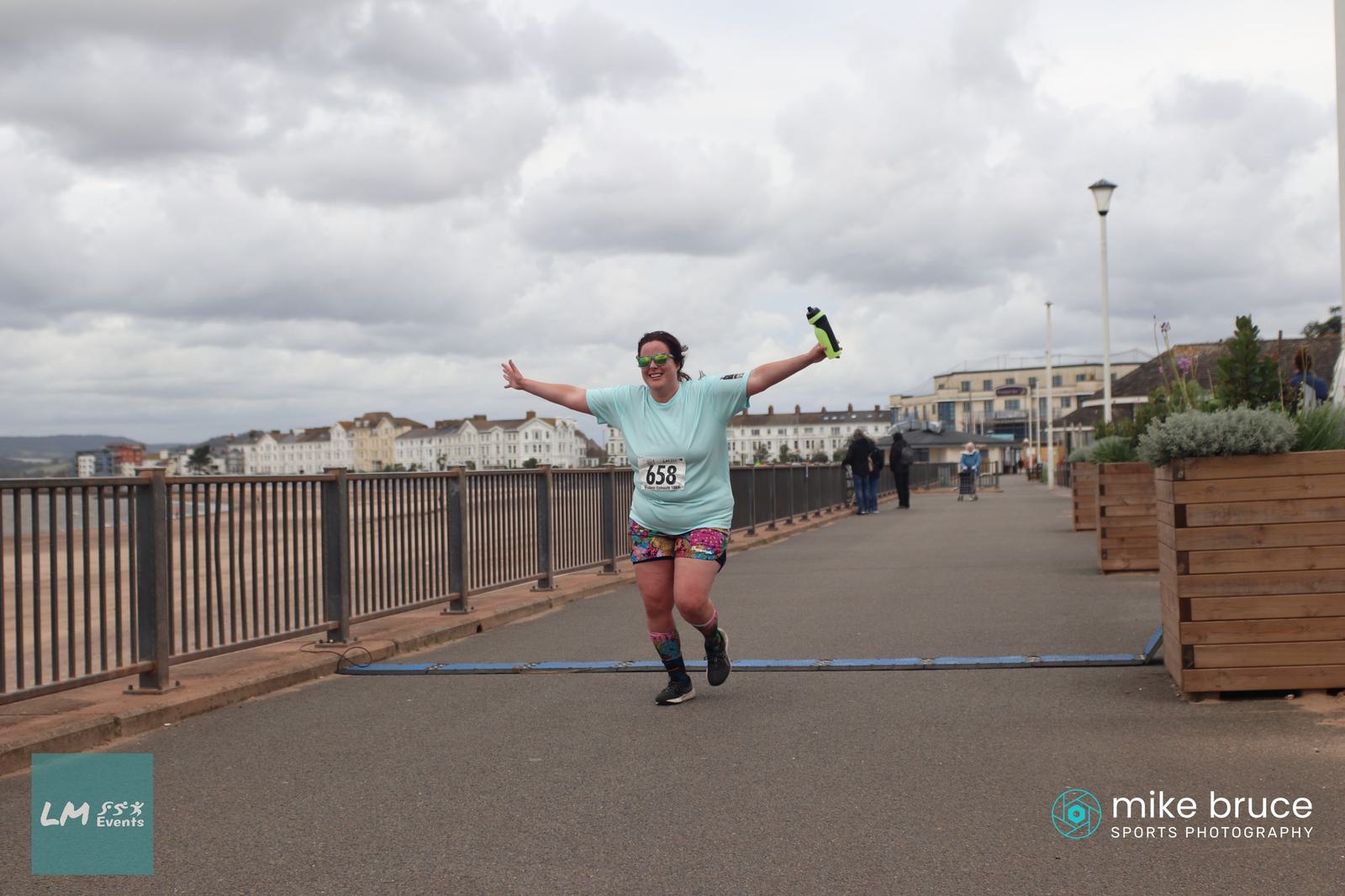 A person wearing a light blue shirt and colorful shorts triumphantly crosses a finish line with arms raised. The background features a boardwalk, buildings, and a cloudy sky. Logos for LM Events and Mike Bruce Sports Photography are visible.
