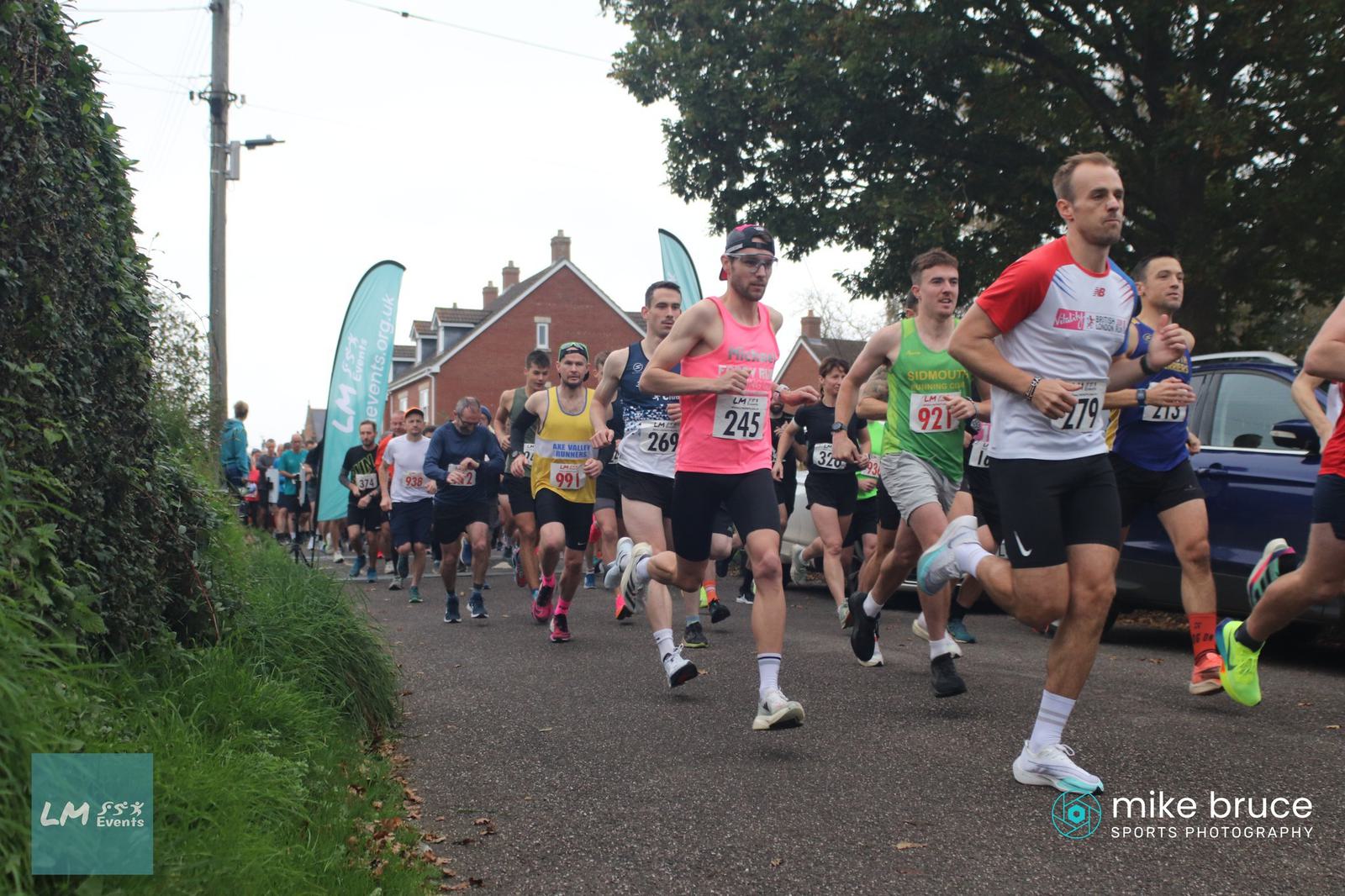A group of runners participating in a race are captured in motion on a tree-lined street. They are wearing assorted athletic gear with numbered bibs. In the background, houses and race banners are visible.