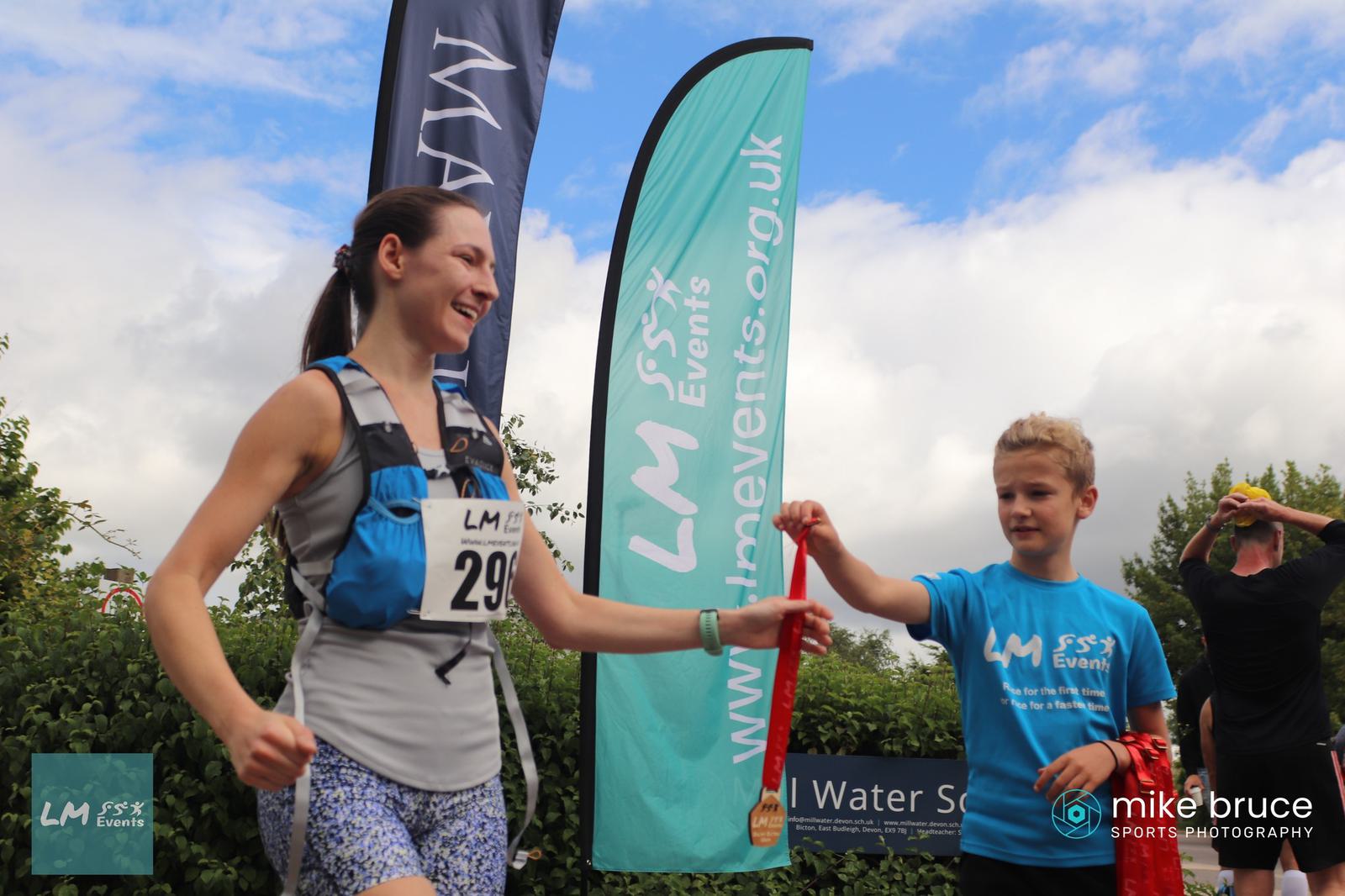 A woman wearing a race bib receives a medal from a young boy at a finish line event. They are smiling. Behind them is a blue flag with "LM Events" text, and the sky is partly cloudy. Another runner is visible in the background.