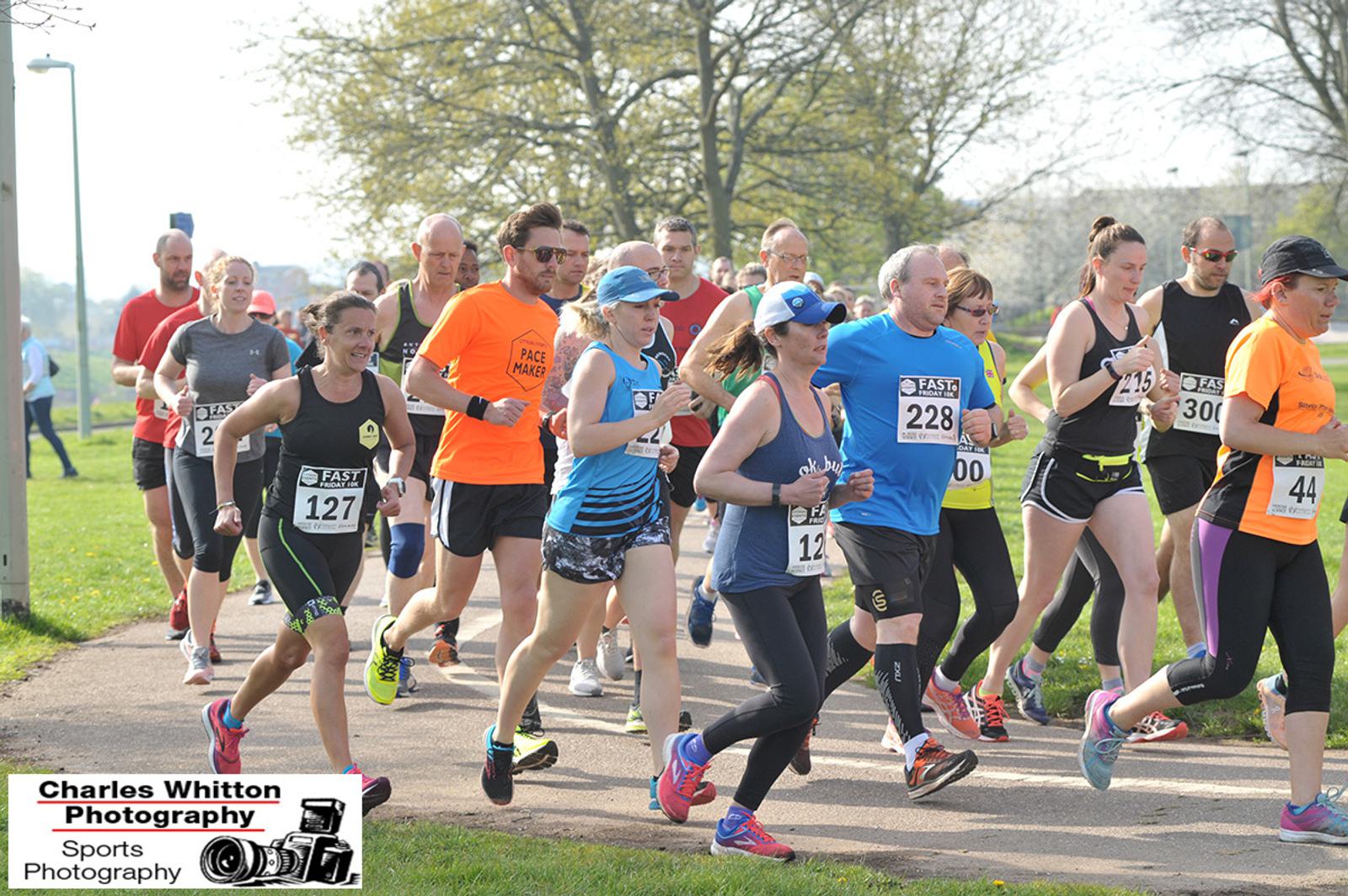 A group of runners participates in a race on a sunny day, surrounded by trees and grass. They wear numbered bibs and various athletic gear. Some participants are in bright colors, with spectators in the background. A photography logo is visible in the corner.