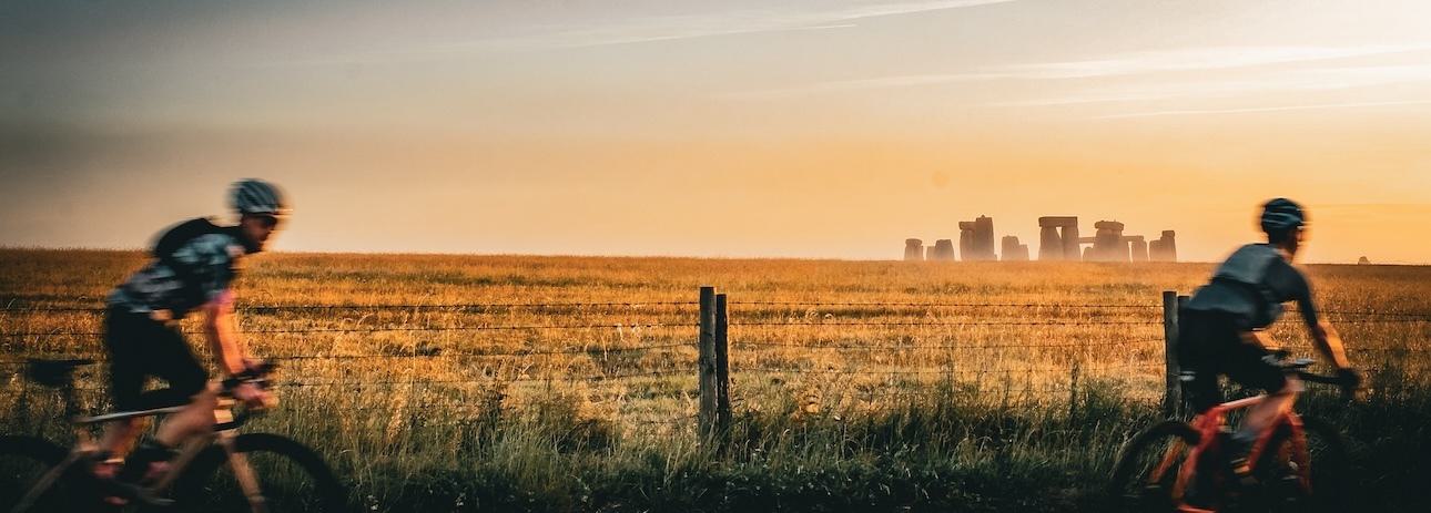 Two cyclists ride past a field at sunset with the silhouette of Stonehenge in the background. The sky is a mix of orange and yellow hues, casting a warm light over the scene.