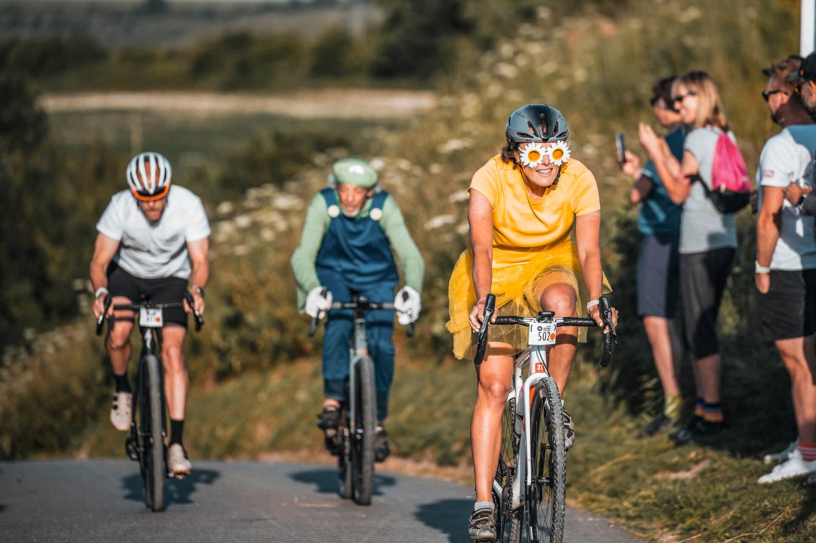 Three cyclists riding on a rural road, with the lead cyclist in a yellow dress wearing sunglasses. Spectators stand by the roadside, some clapping. The background features lush greenery and blurred landscapes under a clear sky.