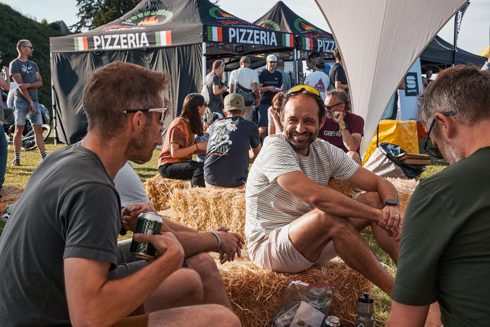 A group of people sit and converse on hay bales at an outdoor event. The background features pizzeria tents and a grassy area. One man in the foreground holds a drink can and wears a striped shirt, smiling at his companions.