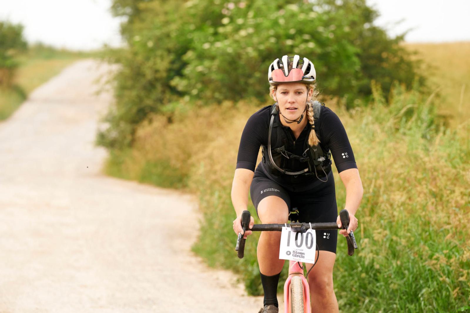 A cyclist wearing a black outfit and helmet rides a gravel road on a bright day, surrounded by greenery. The bike has a race number, and the path stretches into the distance.
