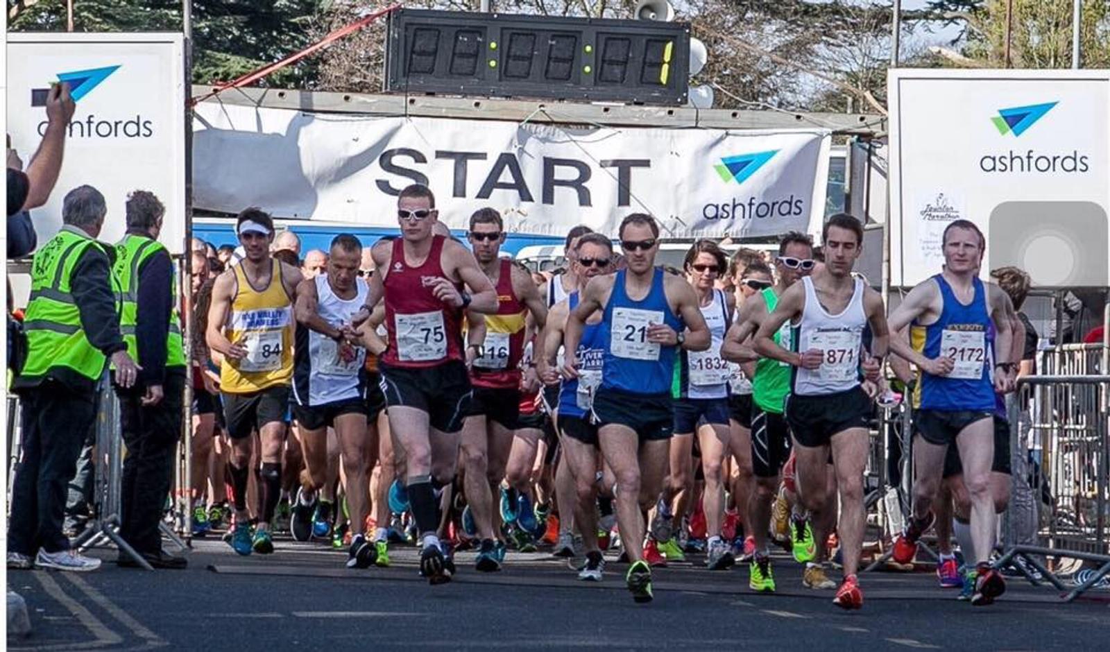 Runners in athletic gear are starting a race at a marked starting line beneath a large clock. Spectators and officials stand nearby. Sunlit trees are visible in the background.