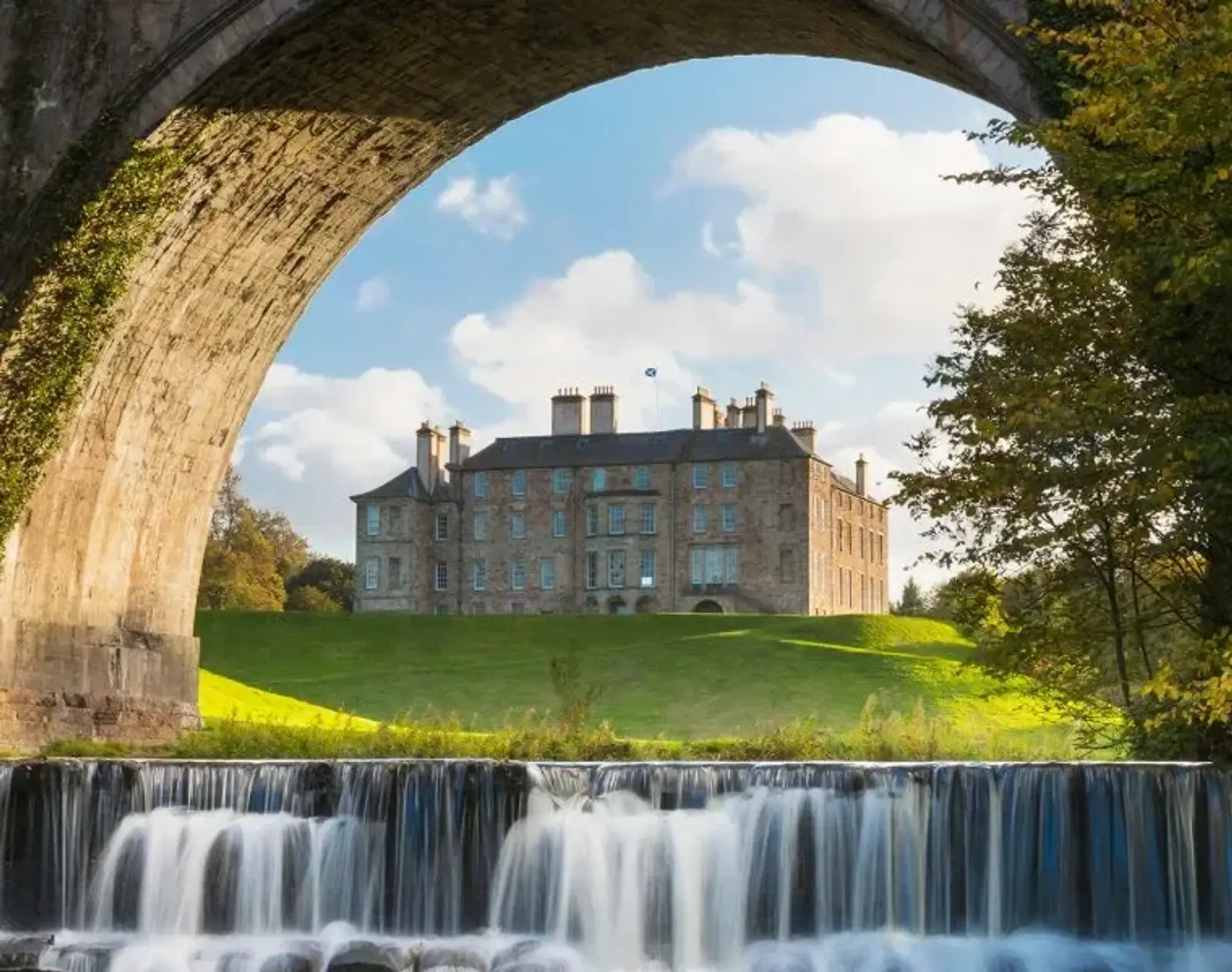 A grand stone mansion is framed beneath a large stone bridge arch. A small waterfall flows in the foreground, set against a lush green lawn. The sky is bright blue with fluffy clouds, enhancing the serene and picturesque landscape.