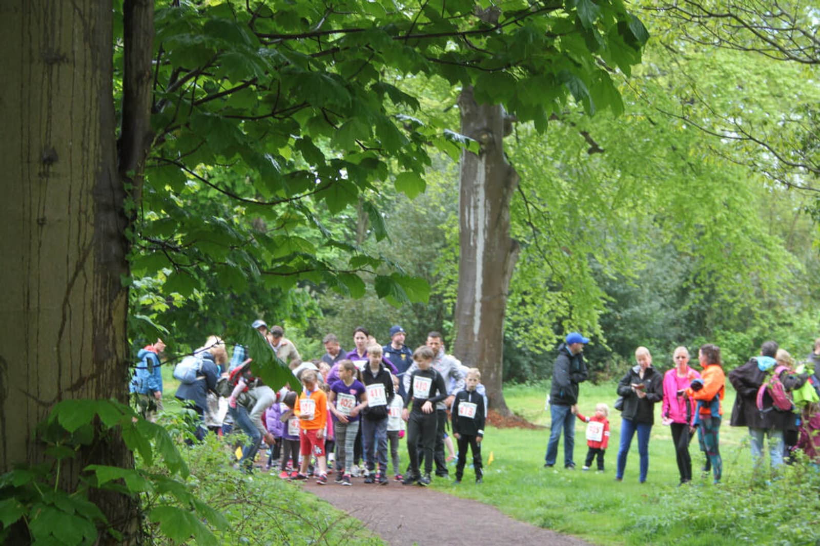 A group of people, including children and adults, gather at the start of a path in a lush, green park. Many wear numbered race bibs, indicating a fun run or race event. Spectators stand nearby, surrounded by tall trees and grass.