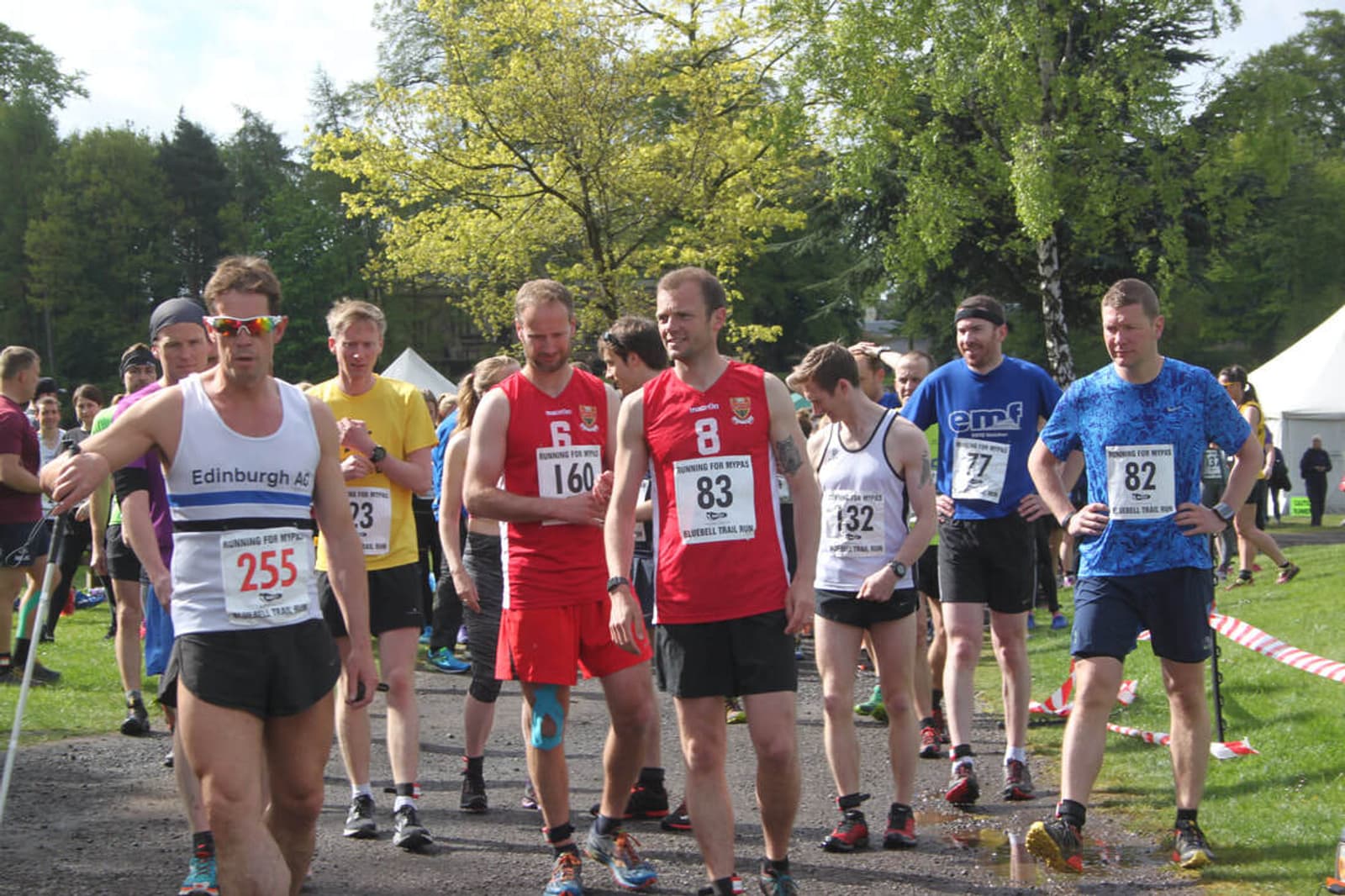 A group of runners wearing race bibs are gathered at the starting line of an outdoor event. They are dressed in athletic gear, and a few trees and a white tent are visible in the background. It's a sunny day.