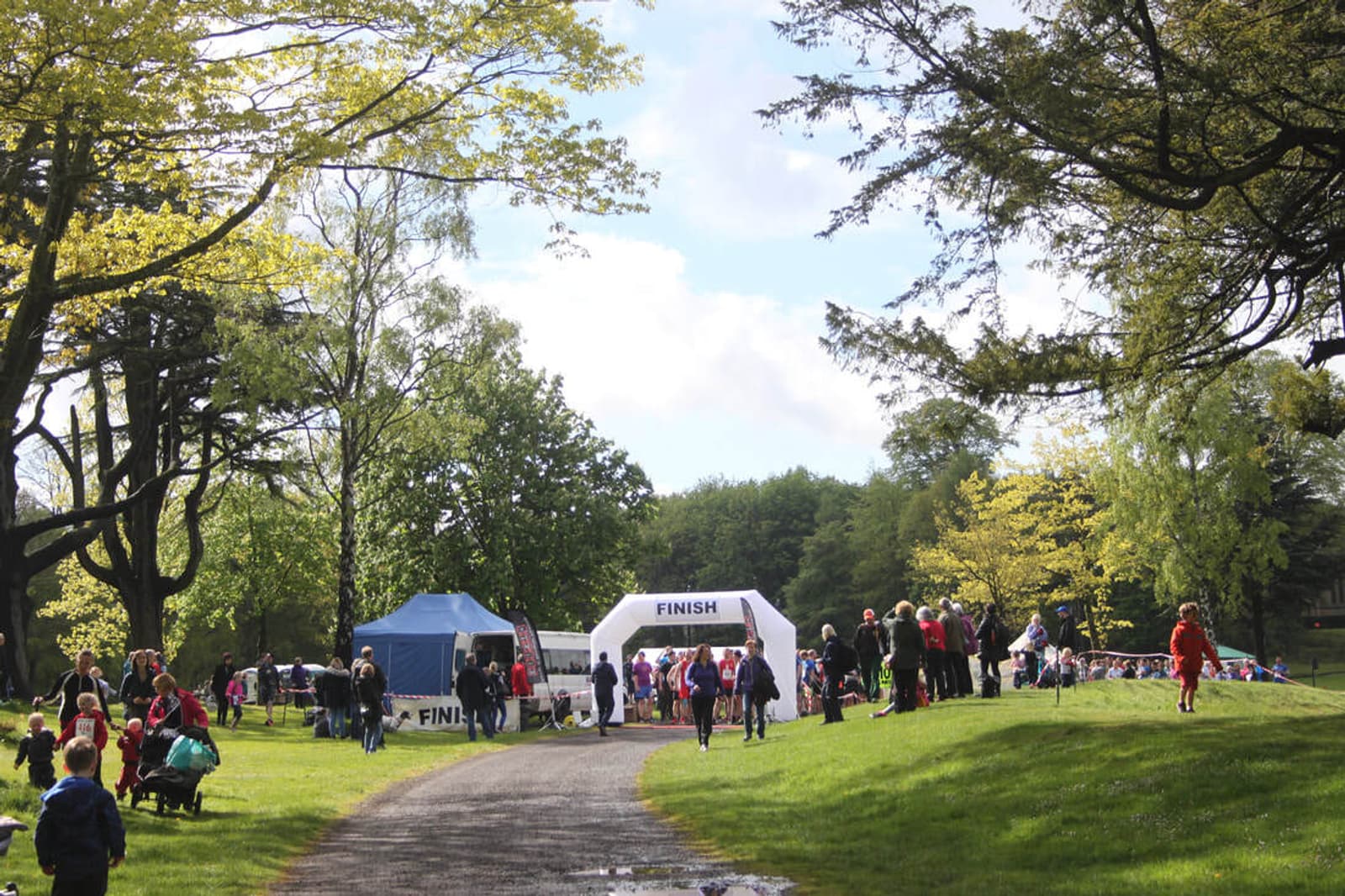A sunny park scene with a crowd gathered around a white inflatable "FINISH" arch. People are walking along a path, children are playing, and trees surround the area. A blue tent is set up nearby.
