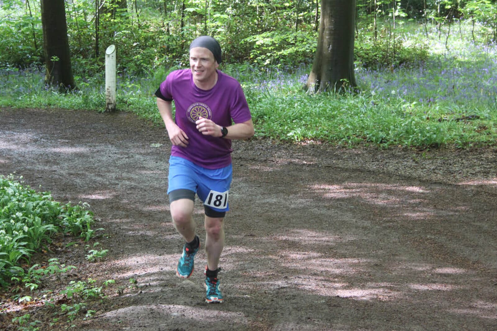 A person is running on a forest trail wearing a purple shirt, blue shorts, and a headband. They have a numbered race bib on their shorts. The background is lush with green foliage and scattered purple flowers.