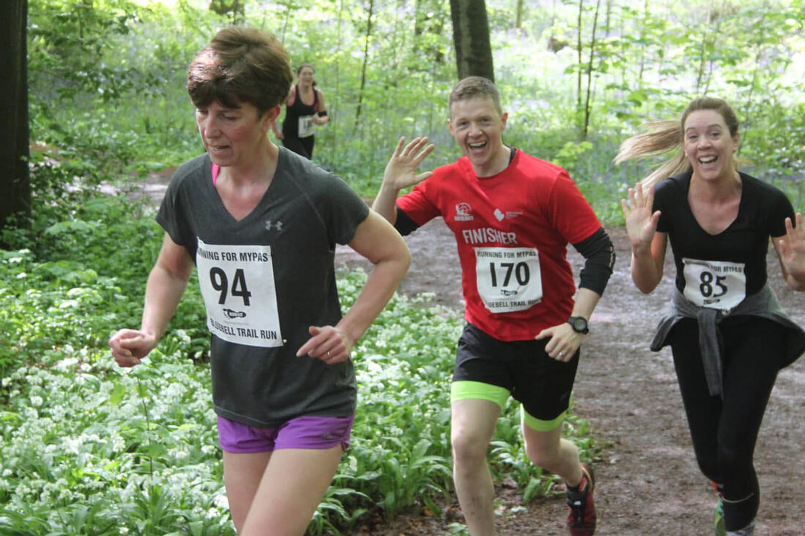 Three people are running on a forest trail. The person in front wears a dark shirt with the number 94. Behind, a person in a red shirt with 170 and another in a black shirt with 85 wave and smile energetically. The trail is surrounded by greenery.