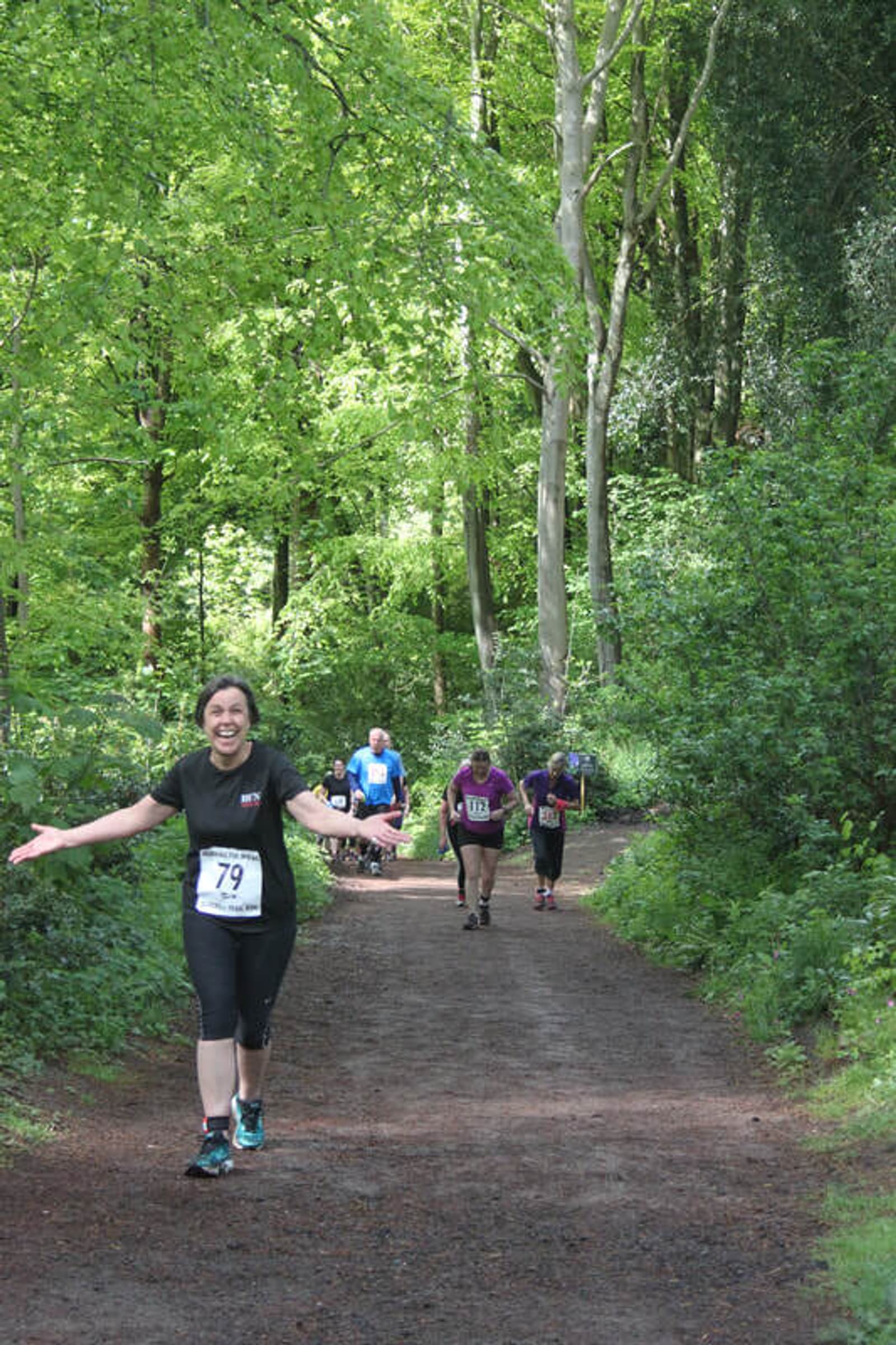 A woman with a race bib number 79 is smiling broadly and spreading her arms wide as she jogs toward the camera on a forest path. Other runners are visible behind her among tall, green trees in a sunlit, leafy forest.