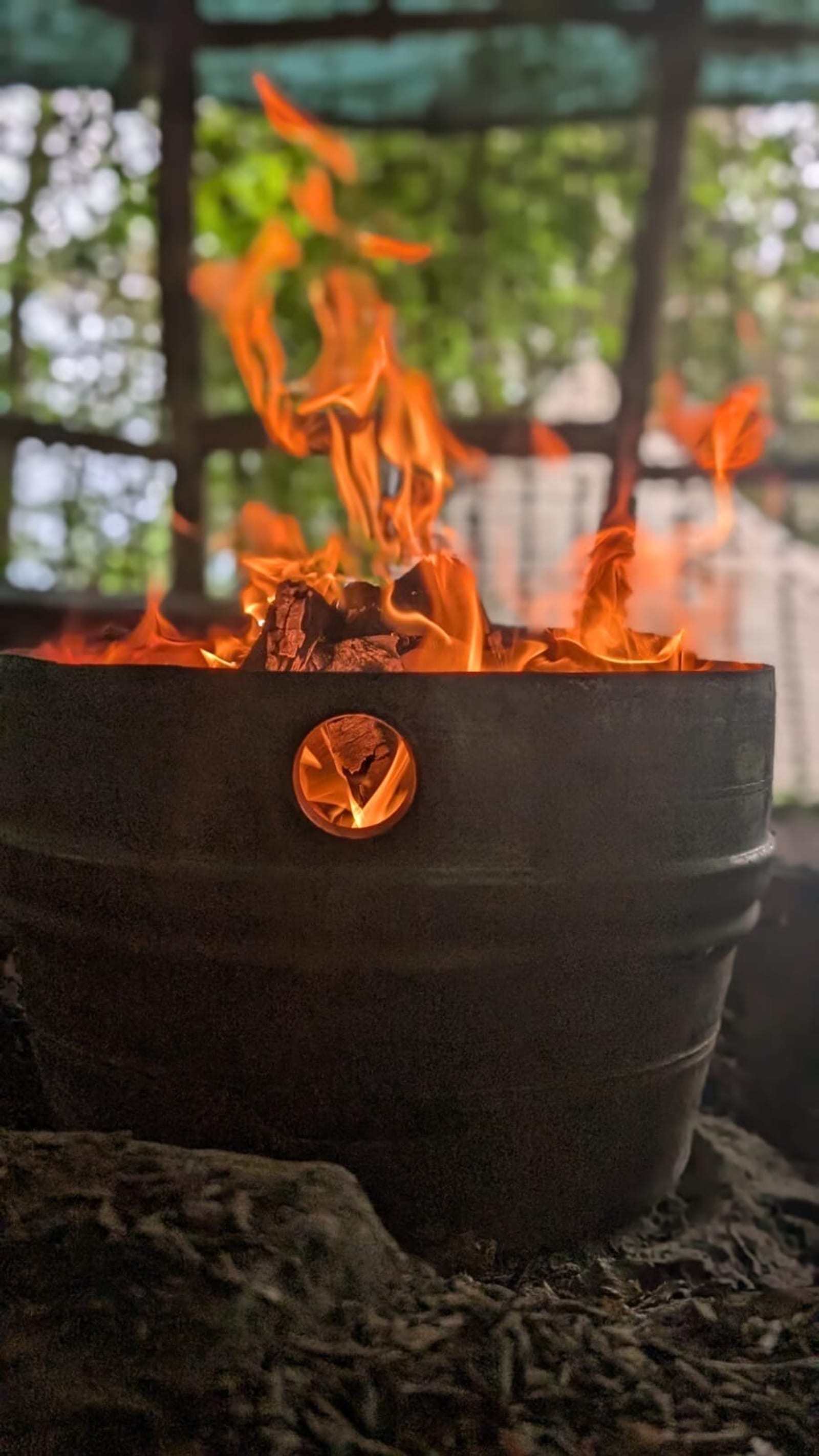A close-up of a metal drum converted into a fire pit, with bright orange flames and burning wood inside. The background shows greenery and a partially blurred outdoor setting.