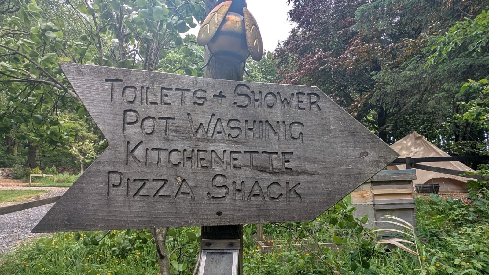 A wooden sign with engraved text points right, listing "Toilets, Shower, Pot Washing, Kitchenette, Pizza Shack." The sign is surrounded by greenery and trees.