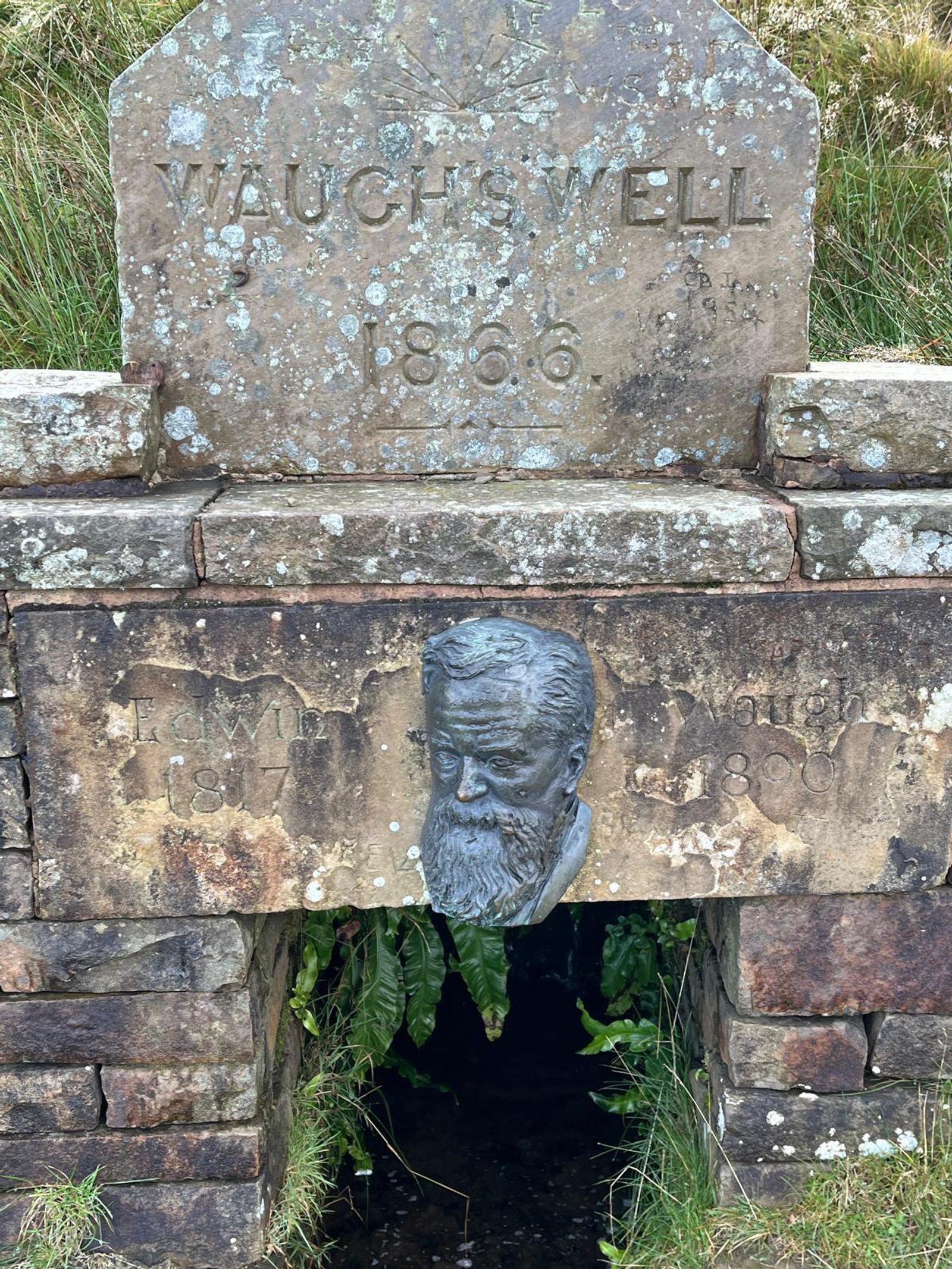 A stone well marked "WAUCH'S WELL 1856" with a carved plaque below showing a bearded man's head and the years 1817–1897, with greenery growing inside the well’s opening beneath.