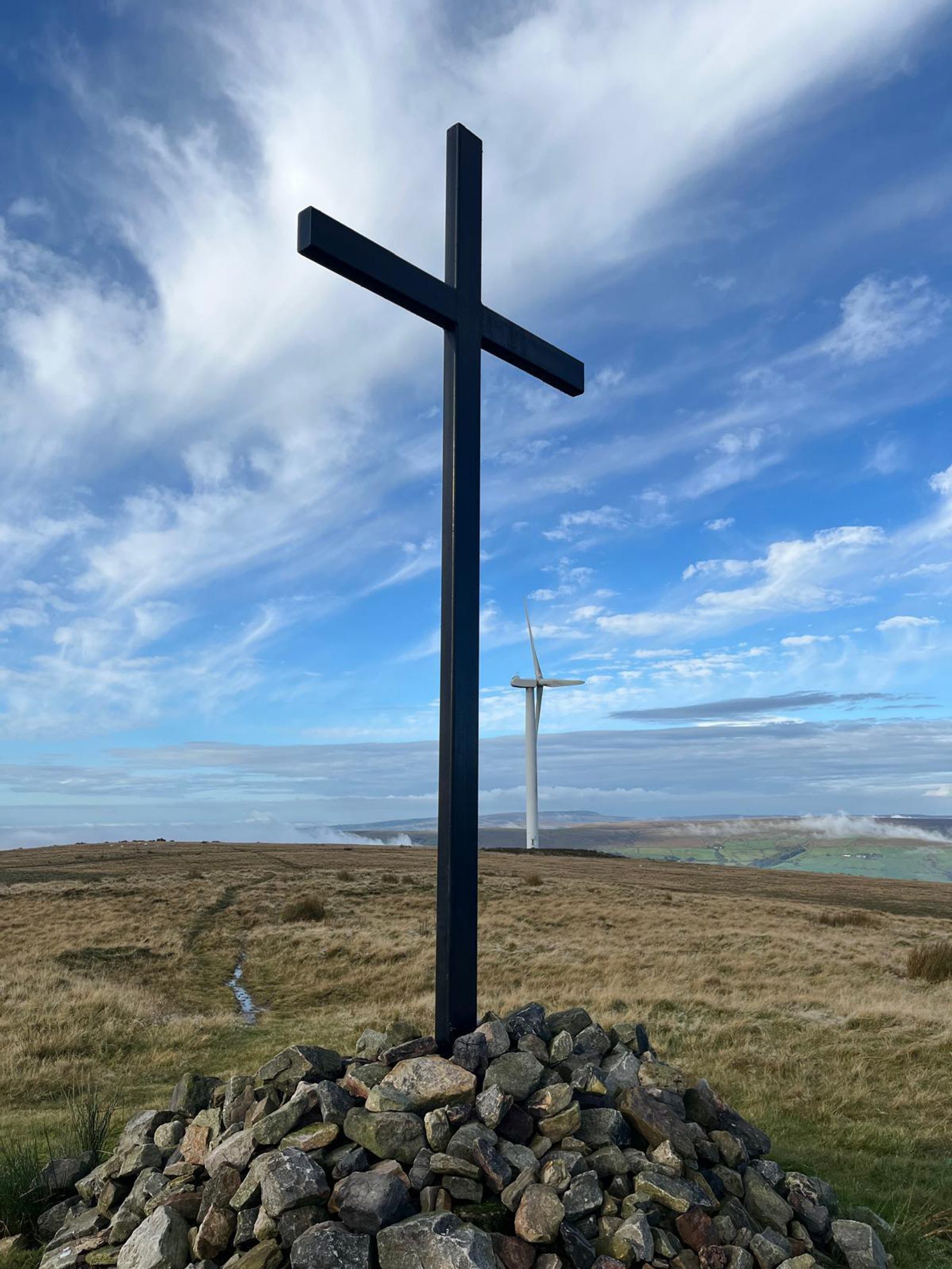 A large black cross stands atop a mound of stones on grassy moorland, with a wind turbine visible in the distance under a blue sky with wispy clouds.