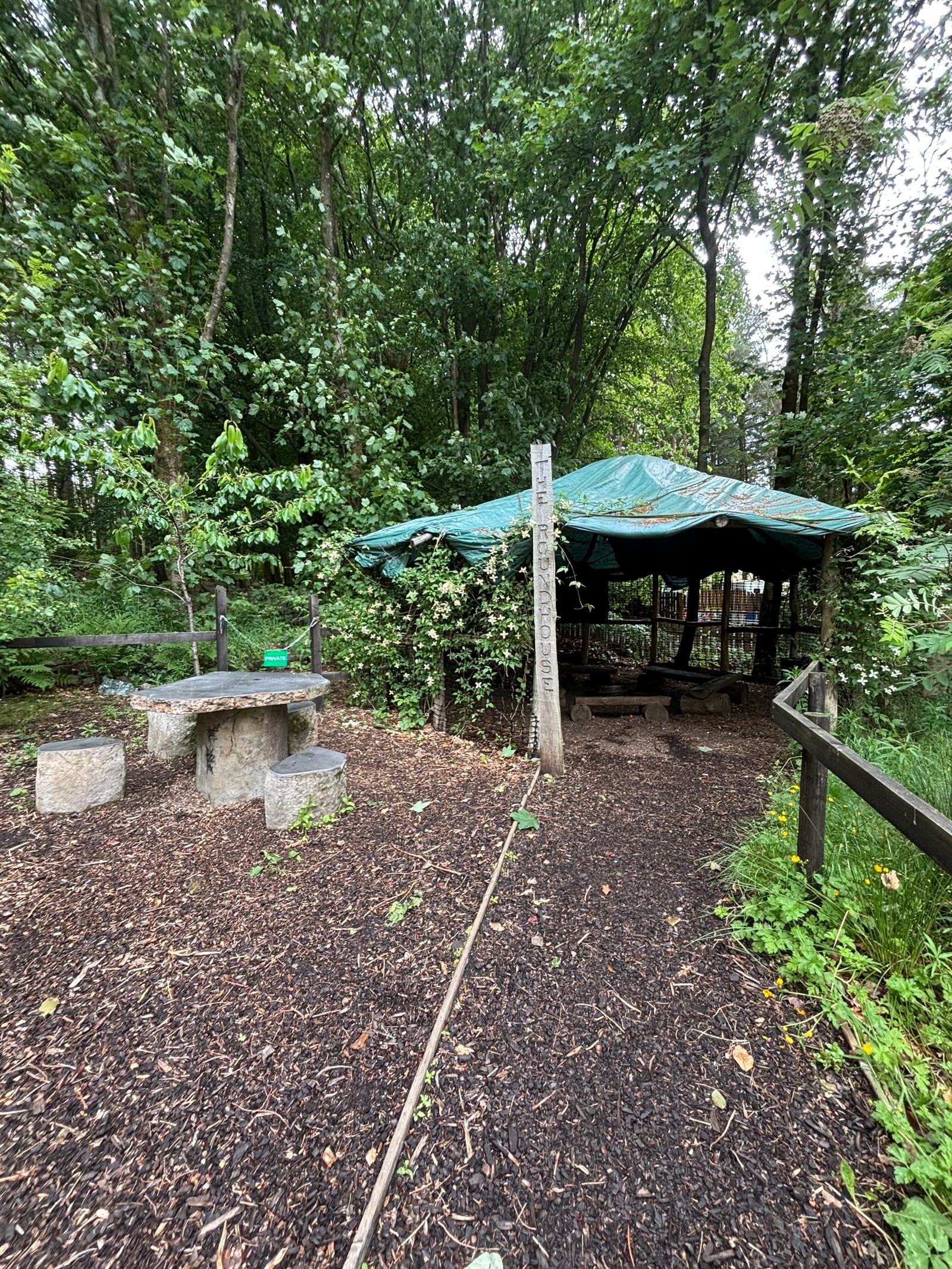 A shaded outdoor area in a wooded setting with a canopy covering benches and tables. Nearby, there is a stone table with stools and a wooden fence along a bark-covered path. Trees and greenery surround the scene.