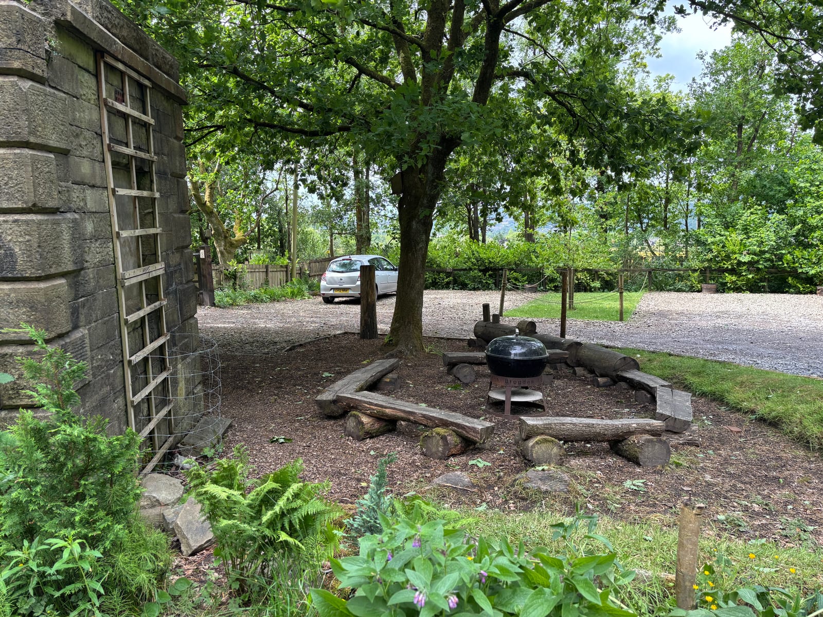 A small fire pit area with a black grill, surrounded by wooden logs for seating, is set under a large tree near a stone building. A silver car is parked on a gravel drive in the background, with greenery all around.