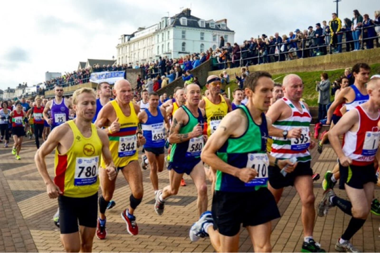 A group of runners in colorful athletic gear participates in a marathon on a brick-paved road. Spectators stand on the sidelines and embankment, cheering them on. A large, white building and cloudy sky are visible in the background.
