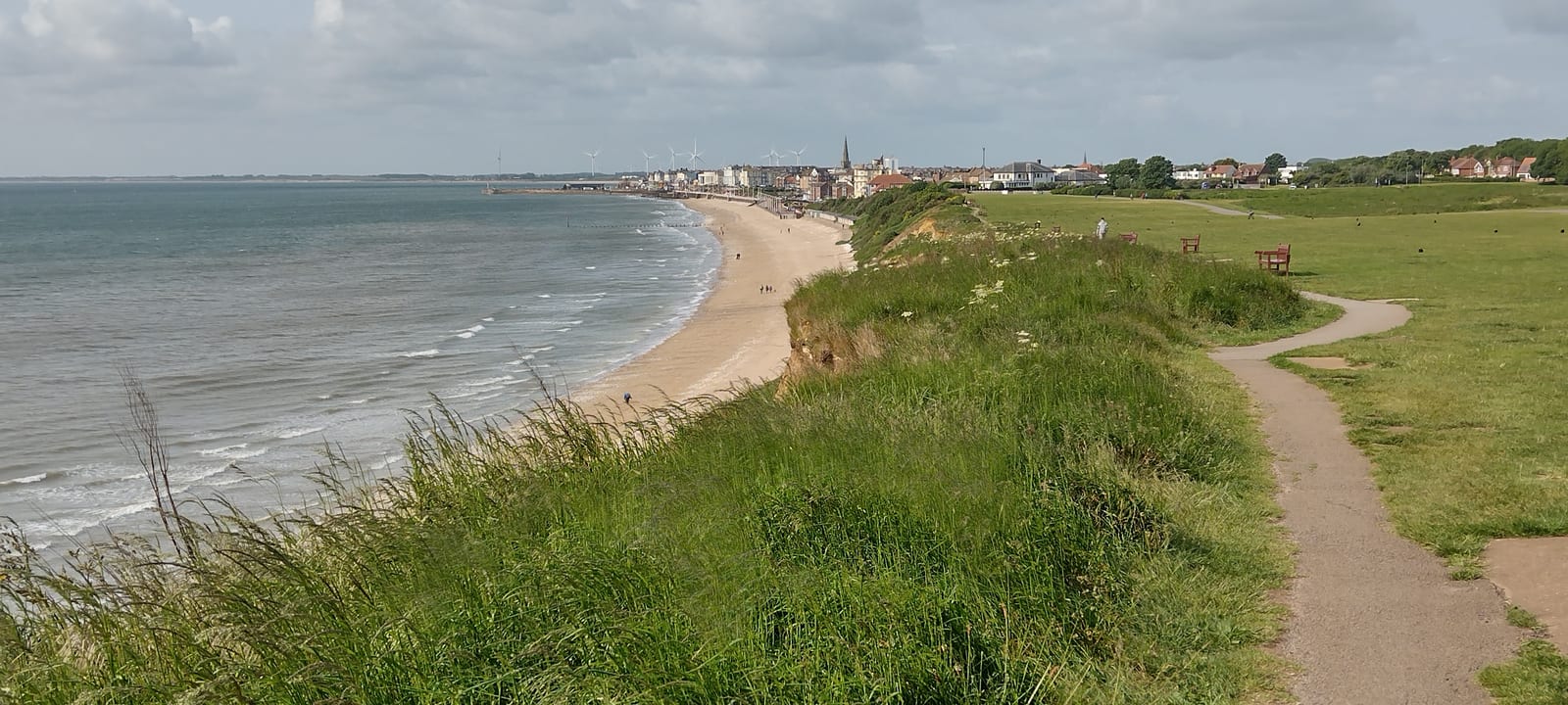 A sandy beach curves along the coastline beside a grassy cliff, with a paved path and benches. Buildings and wind turbines are visible in the distance under a partly cloudy sky.