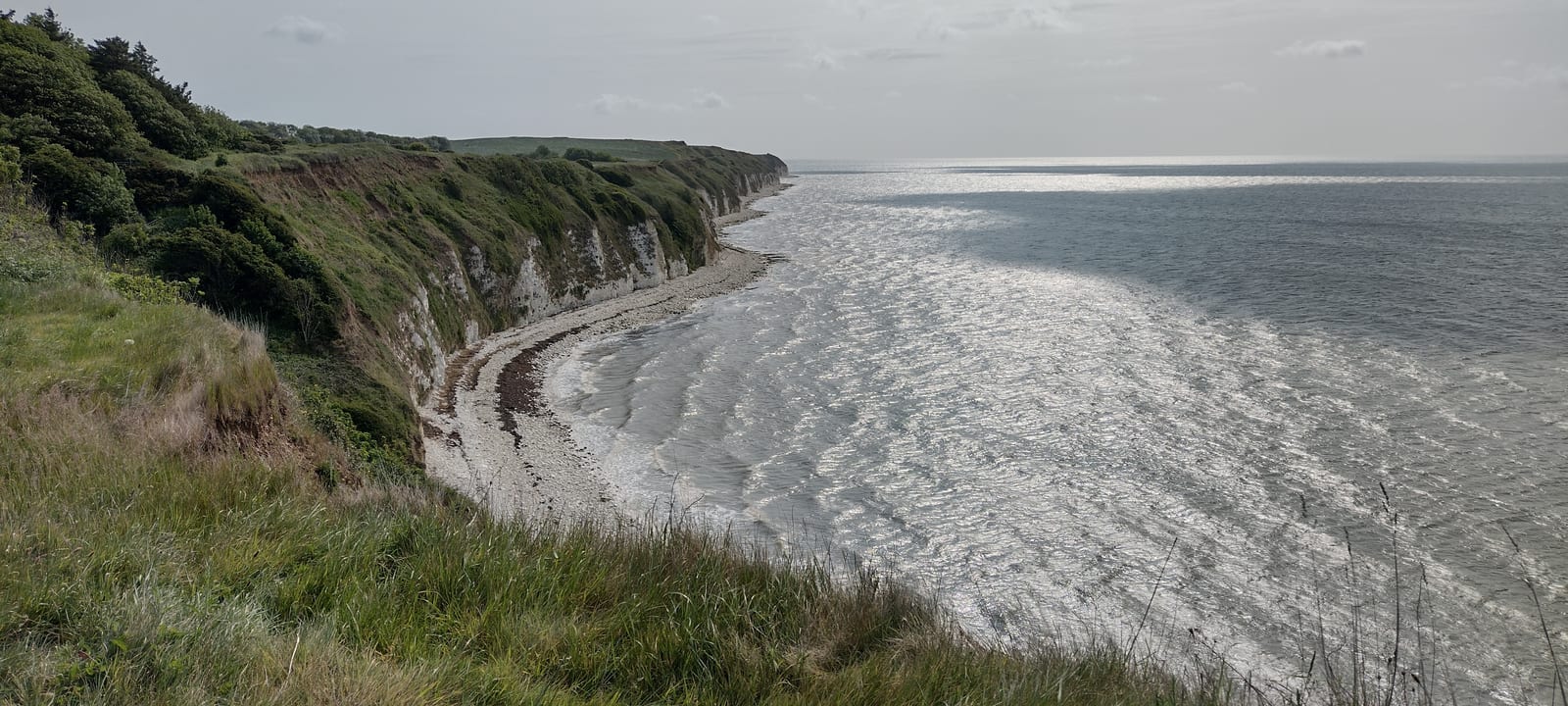 A high coastal cliff covered in green grass overlooks a curved shoreline with white pebbles and gentle waves under a cloudy sky. The sea stretches to the horizon on a calm day.