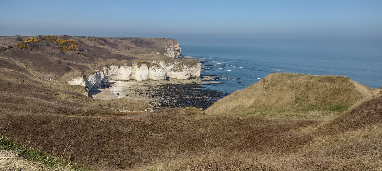 Cliffs overlooking the sea, with white limestone formations, rocky shoreline, and grassy hills in the foreground under a clear blue sky.